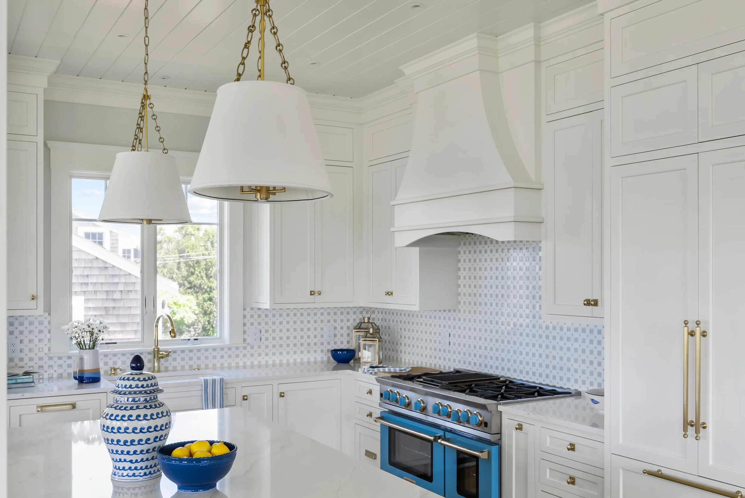 Bright white kitchen with large island, gold hardware, blue accents, and a black and gold oven. Decor includes a blue bowl of lemons and white flowers.