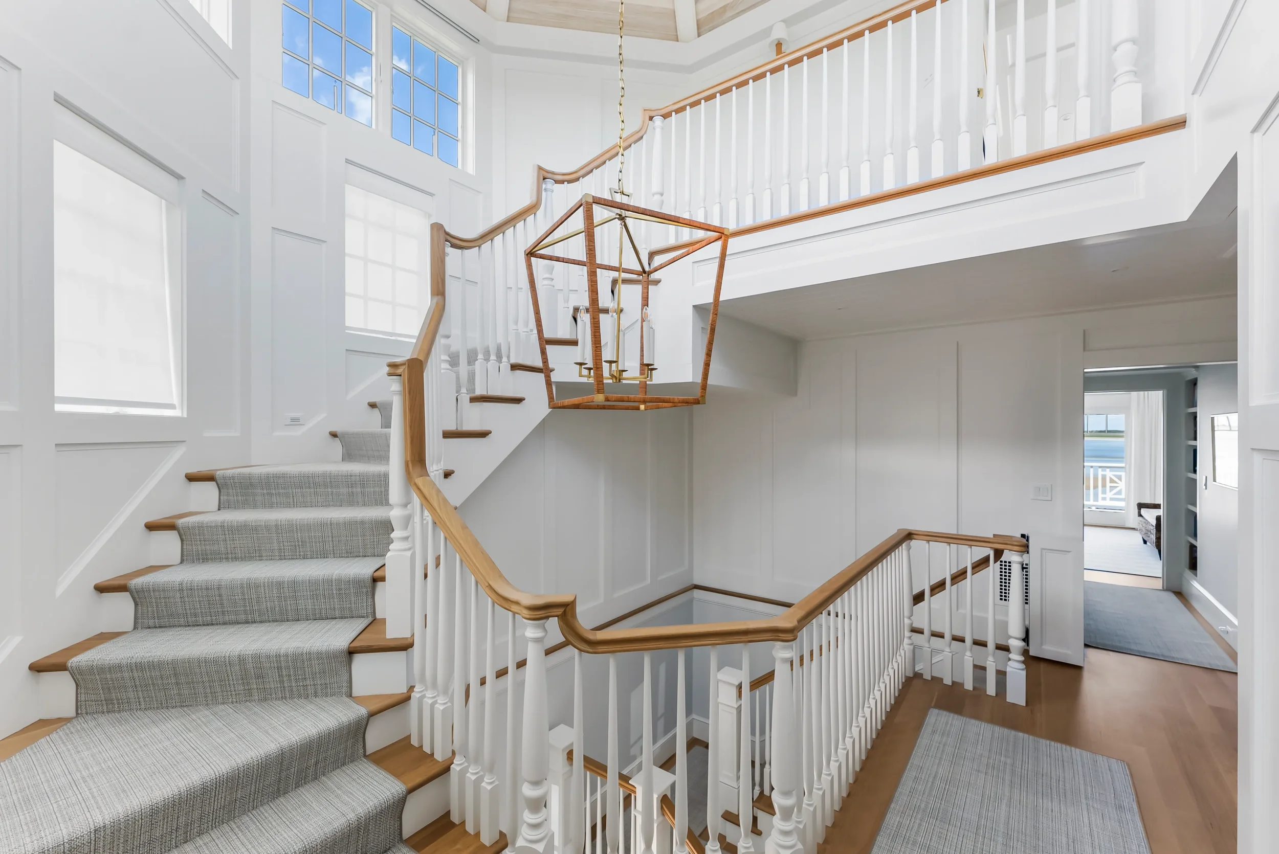 Interior view of a stairway with white wood railings and light wood steps, with a hanging geometric chandelier, leading to a second floor with a balcony overlooking the lower level. Large windows let in natural light.