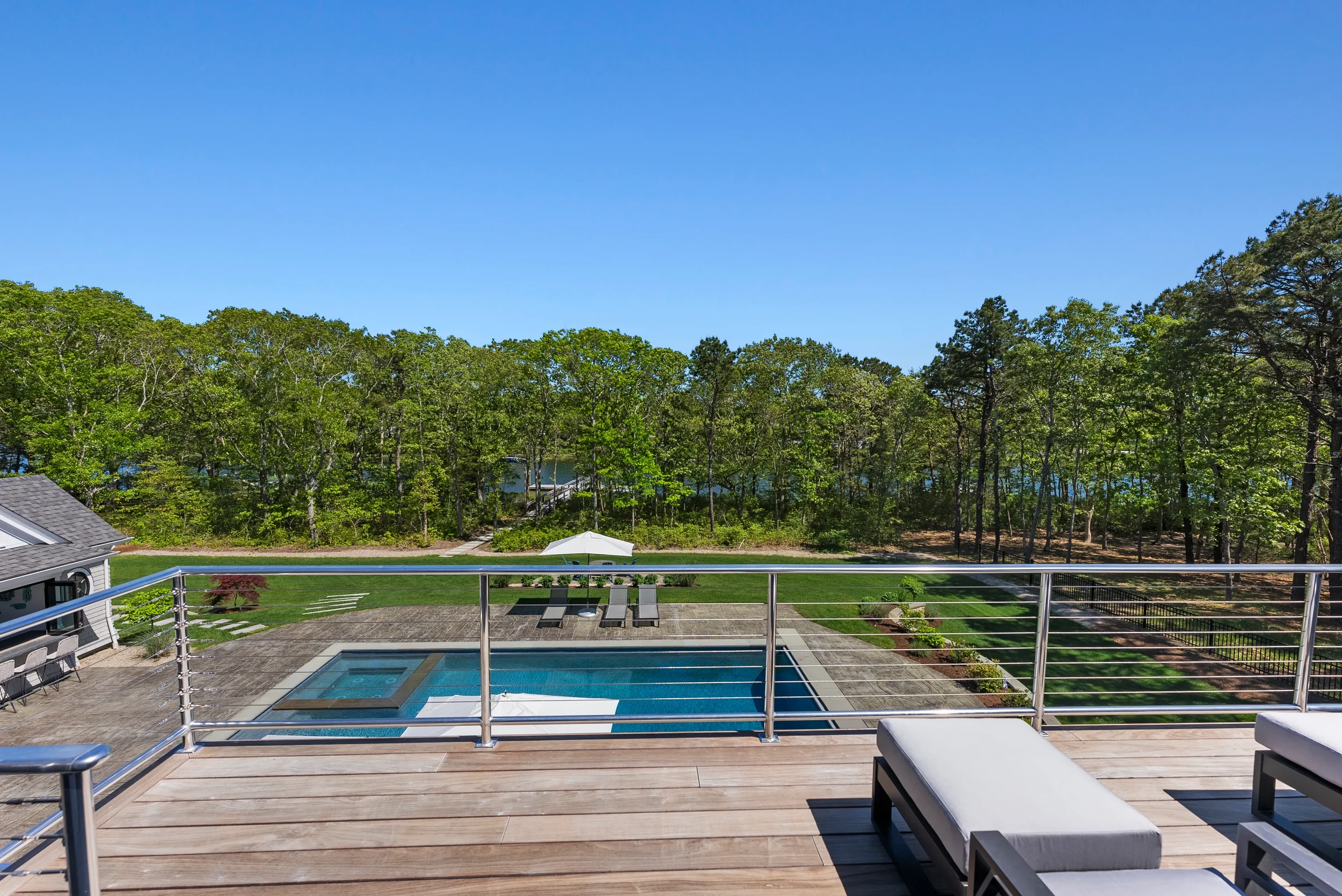 View from a balcony overlooking a backyard with a swimming pool, lounge chairs with umbrellas, manicured lawn, and a dense forest of trees under a clear blue sky.
