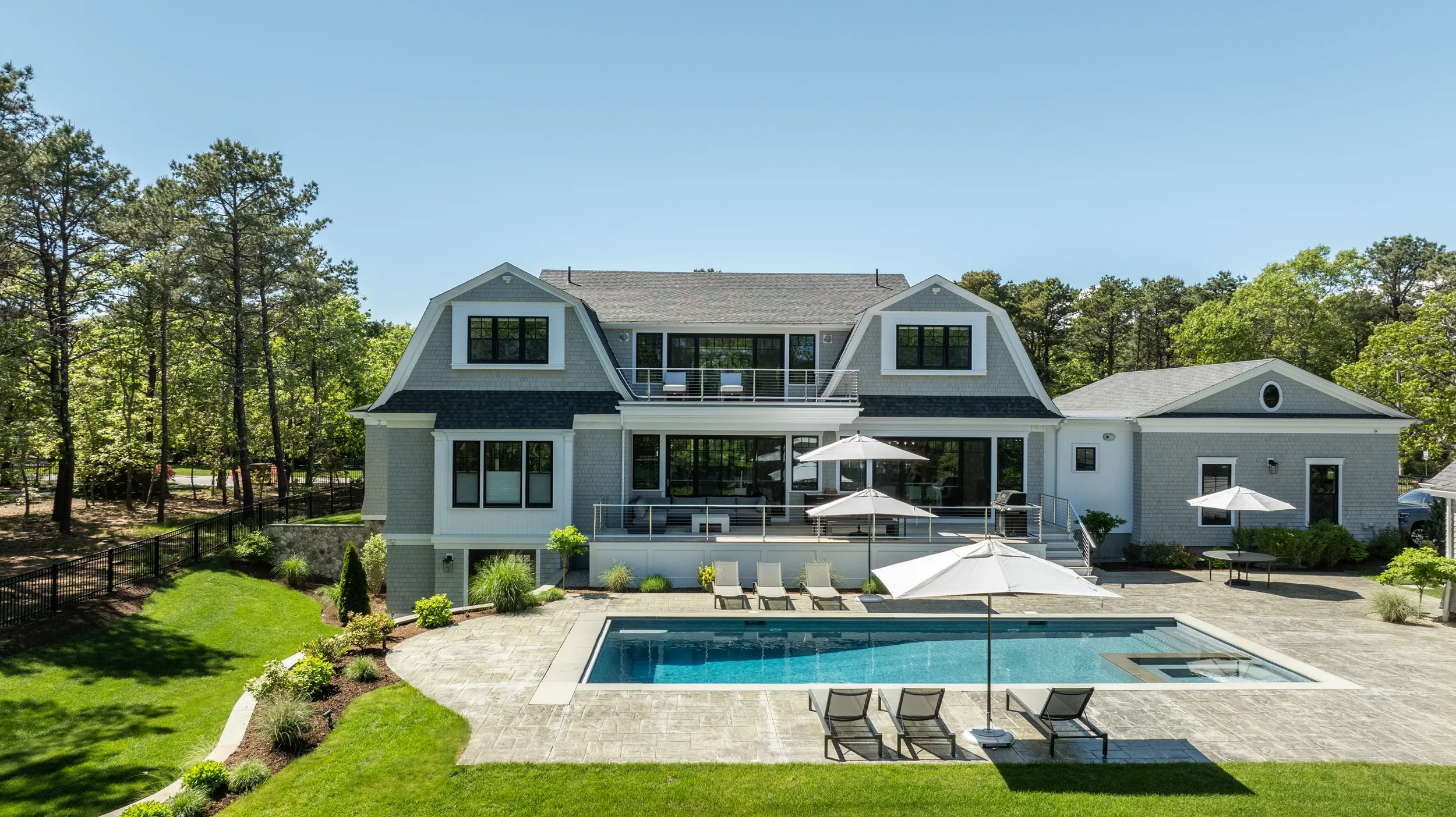 A large modern house with a backyard swimming pool, surrounded by green grass, trees, and outdoor furniture with umbrellas under a clear blue sky.