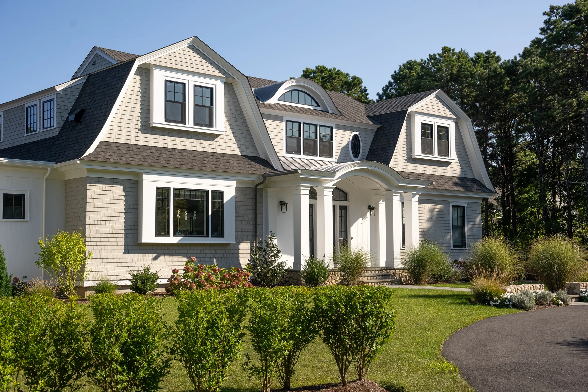 Large, modern house with gray and white exterior, multiple windows, a curved porch, a well-maintained lawn, and surrounded by trees.