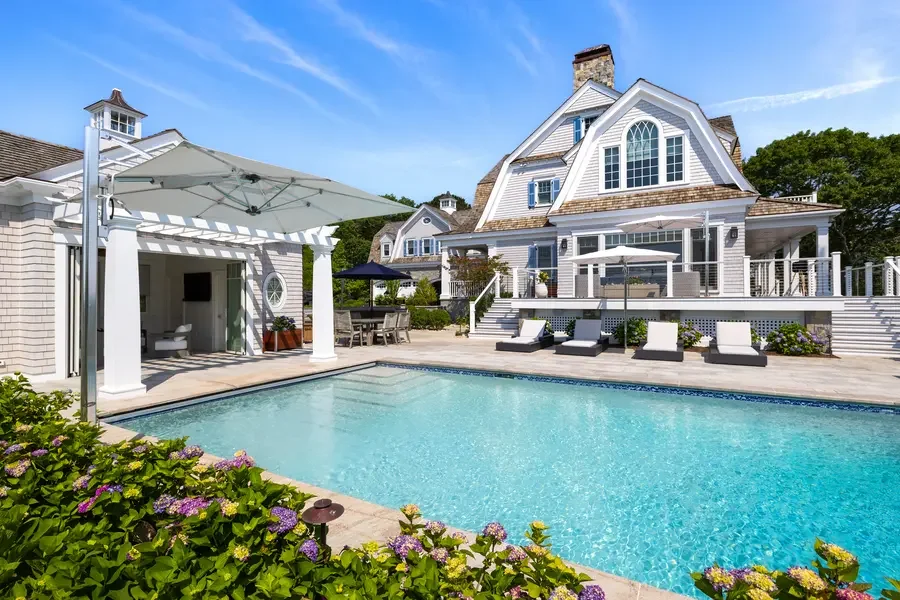 Beautiful backyard with a swimming pool, lounge chairs, umbrellas, and a large white house with a deck, surrounded by greenery and blooming plants.