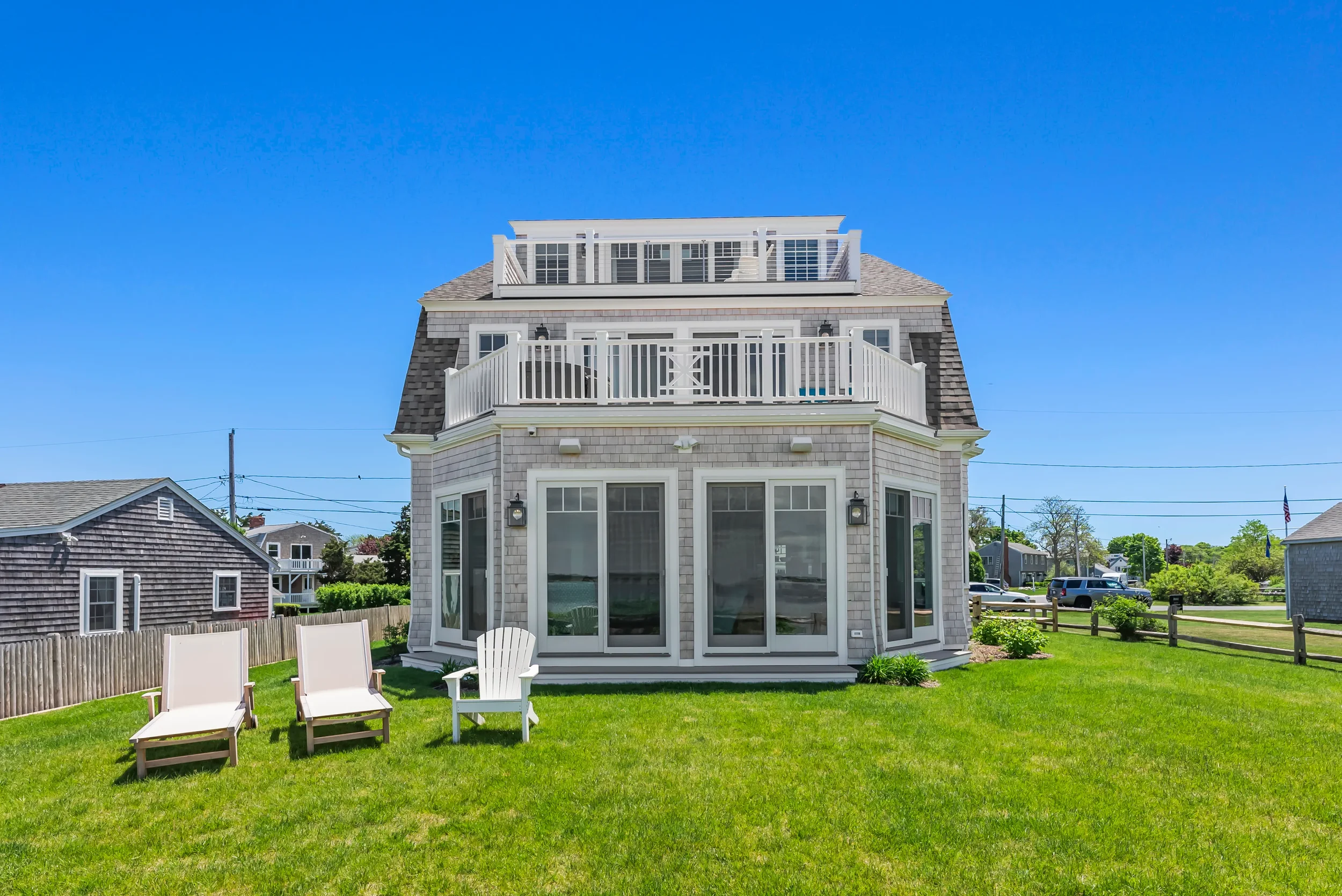Backyard view of a house with large windows, multiple balconies, and outdoor furniture on a grassy lawn, with neighboring houses and a clear blue sky in the background.
