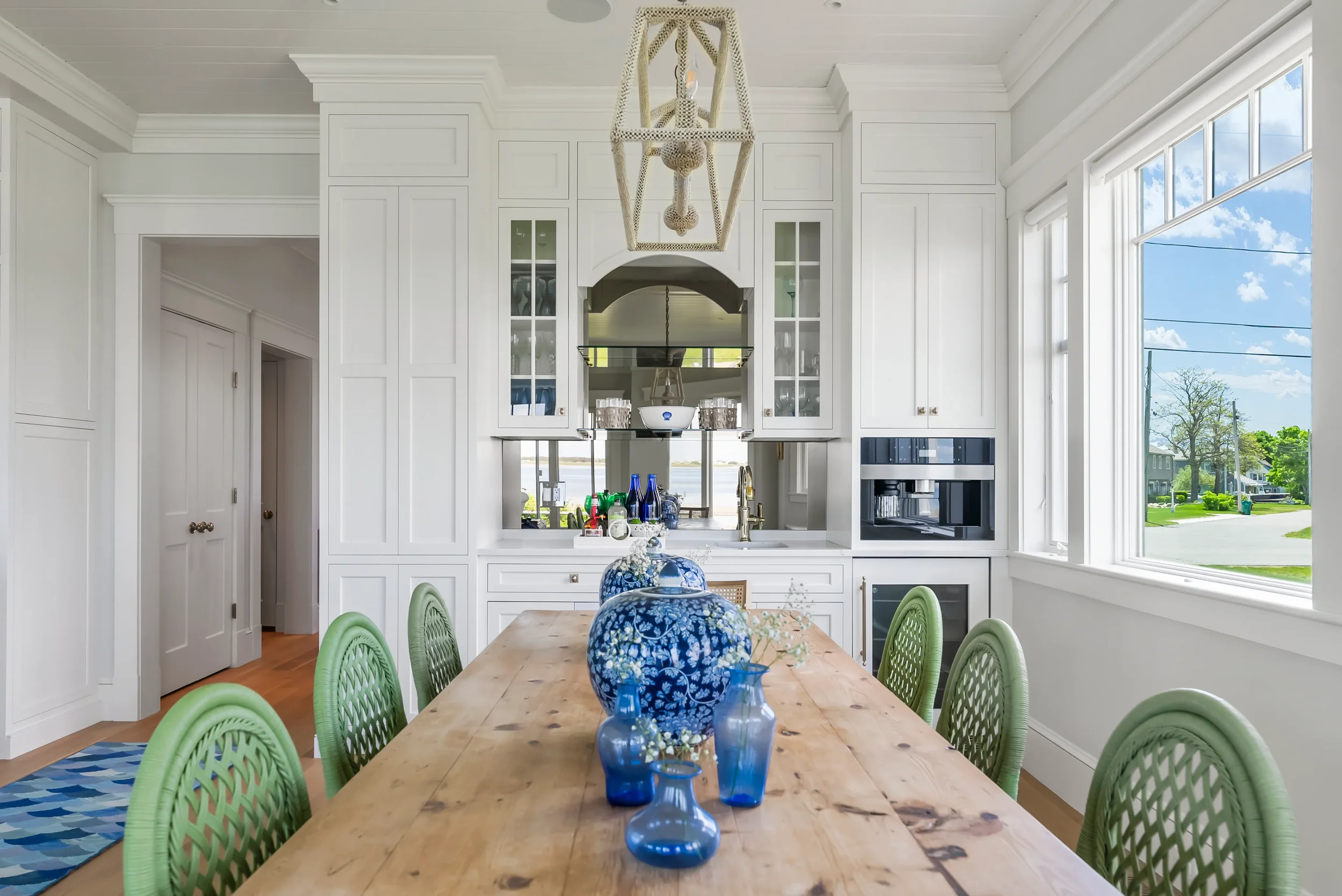 A bright dining room with a wooden table, railing chairs, and a white built-in kitchen cabinet with glass doors. There are blue vases with white flowers and a large blue decorative jar on the table. Large windows on the side reveal a sunny neighborho