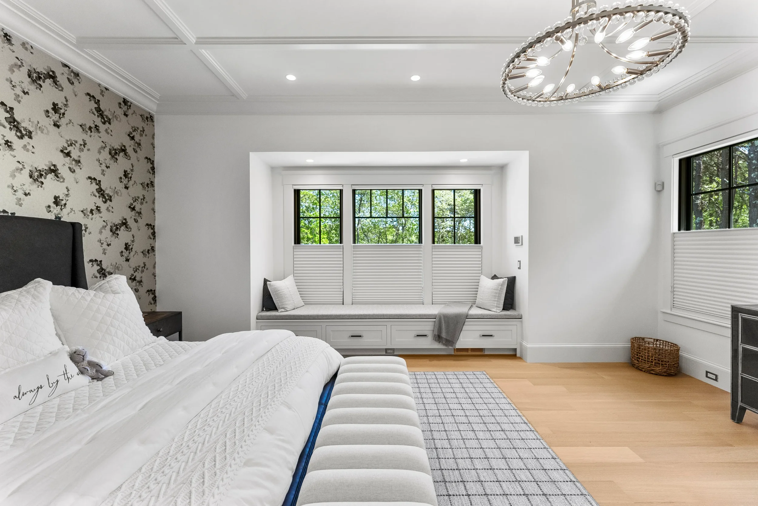 Bright bedroom with a large bed with white bedding, a built-in window seat with cushions, a modern chandelier, and light wood floors.