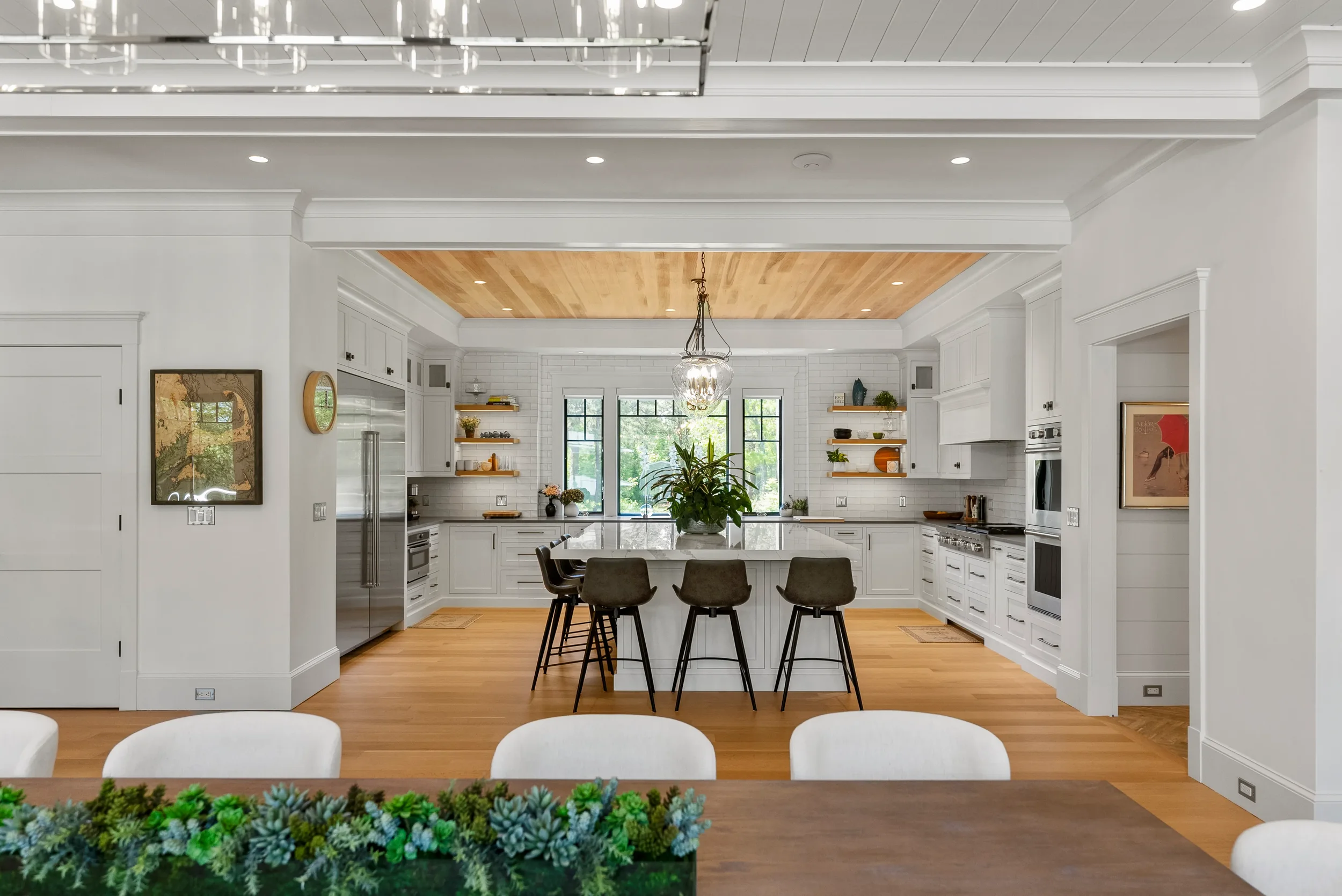 Modern kitchen with white cabinetry, a large island with barstools, and a wooden ceiling detail. Large windows at the back let in natural light, and there are open shelves with decorative items.