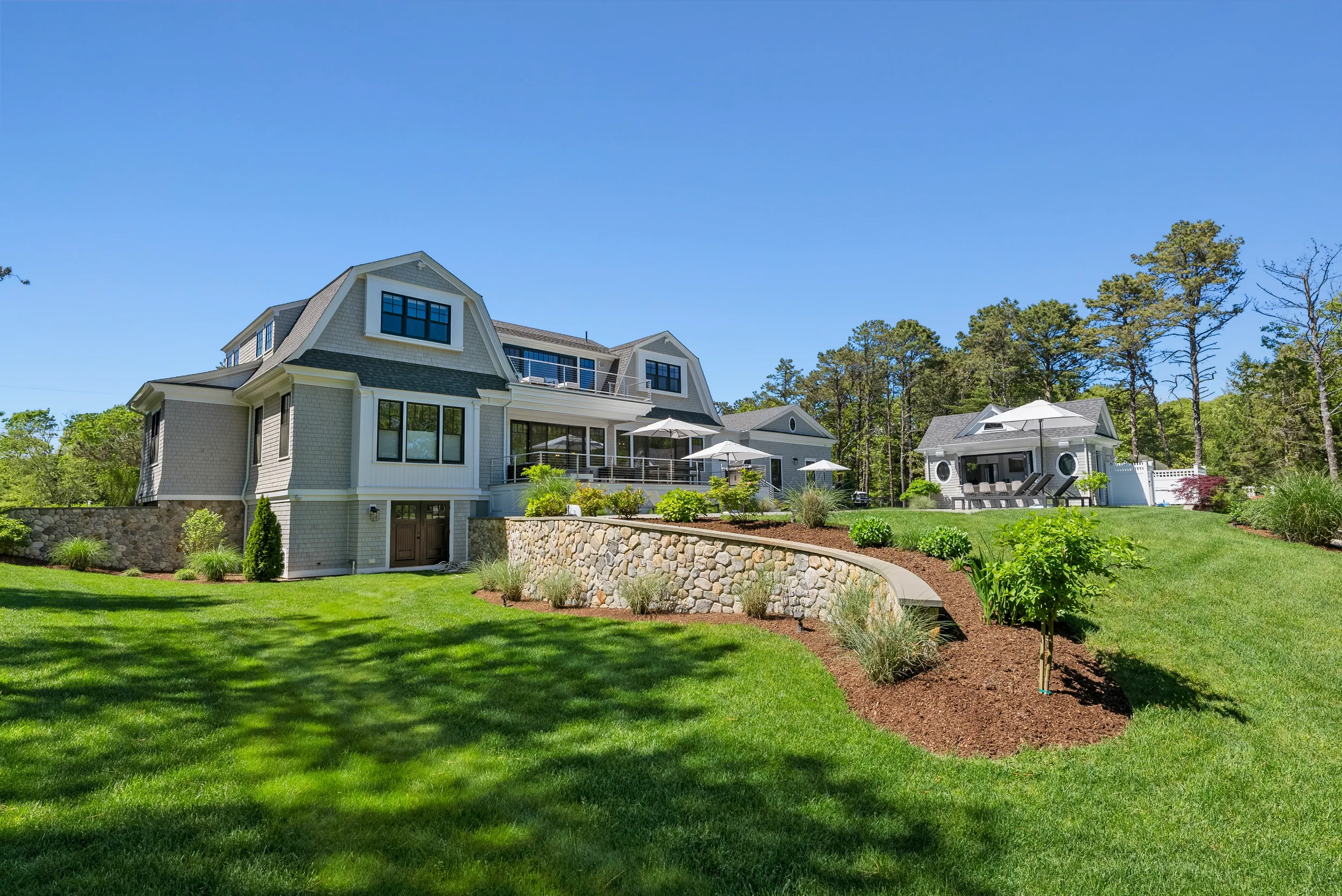 A large multi-story house surrounded by a well-maintained lawn and garden, with a small detached building, trees in the background, and blue sky overhead.