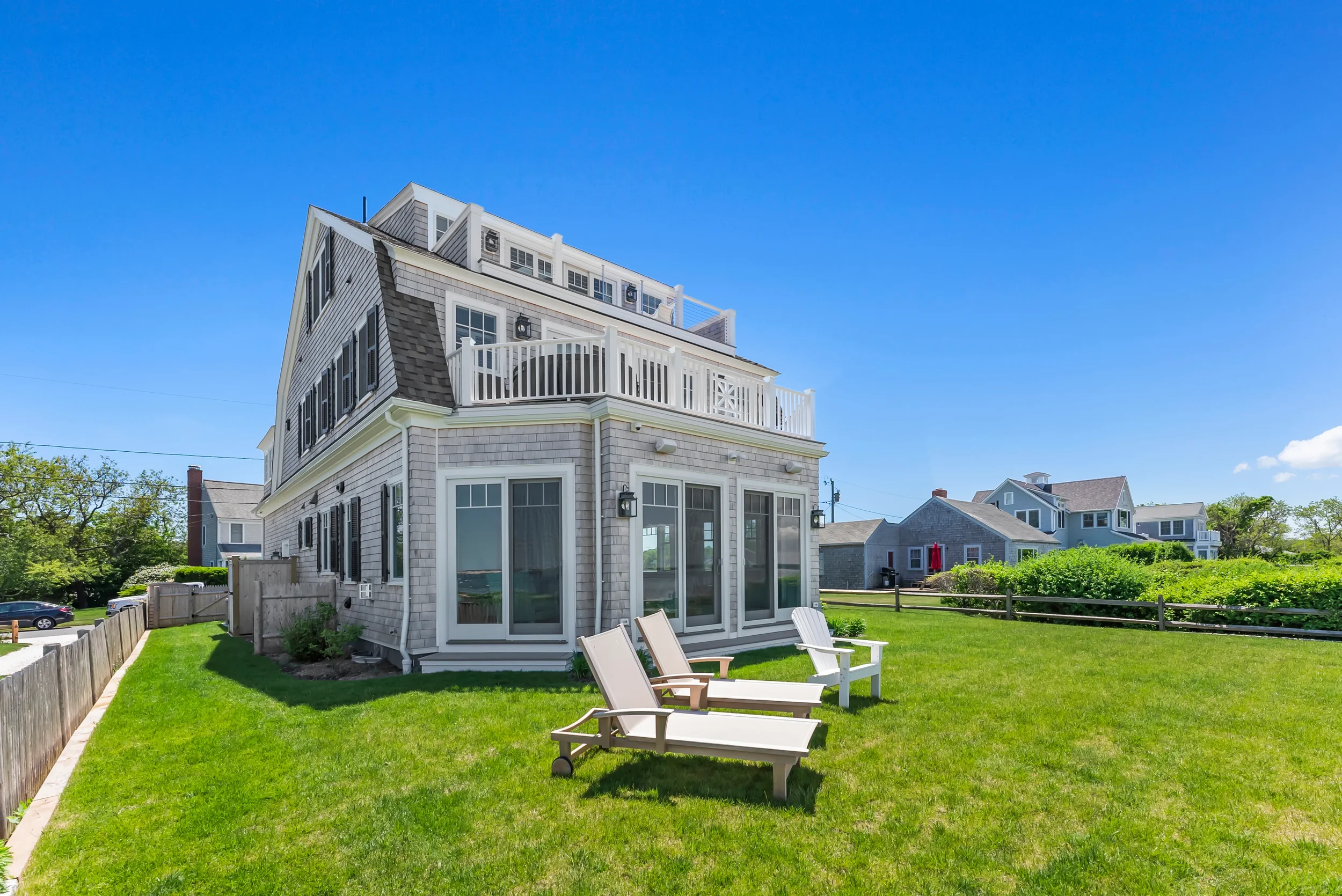 Beach house with multiple levels, large windows, and outdoor deck with white railing, surrounded by green lawn and Adirondack chairs, under a bright blue sky.