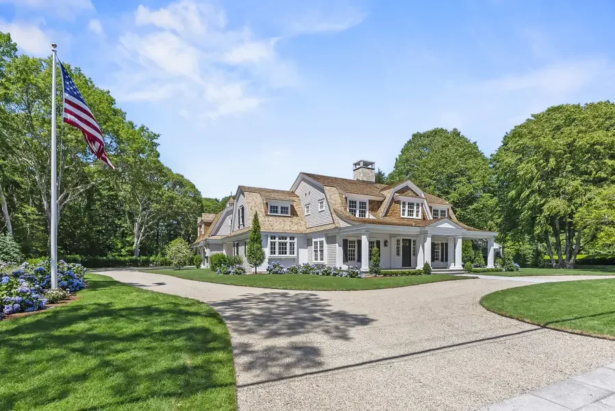 This image showcases a stunning Cape Cod-style estate home set against a brilliant blue sky. The expansive residence features classic New England architecture with weathered cedar shingle siding in natural tones and white trim throughout. The complex