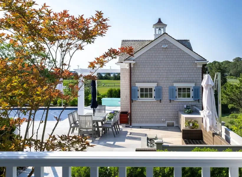 Outdoor backyard patio with a dining table, chairs, umbrellas, a swimming pool, and a grey house with blue shutters.