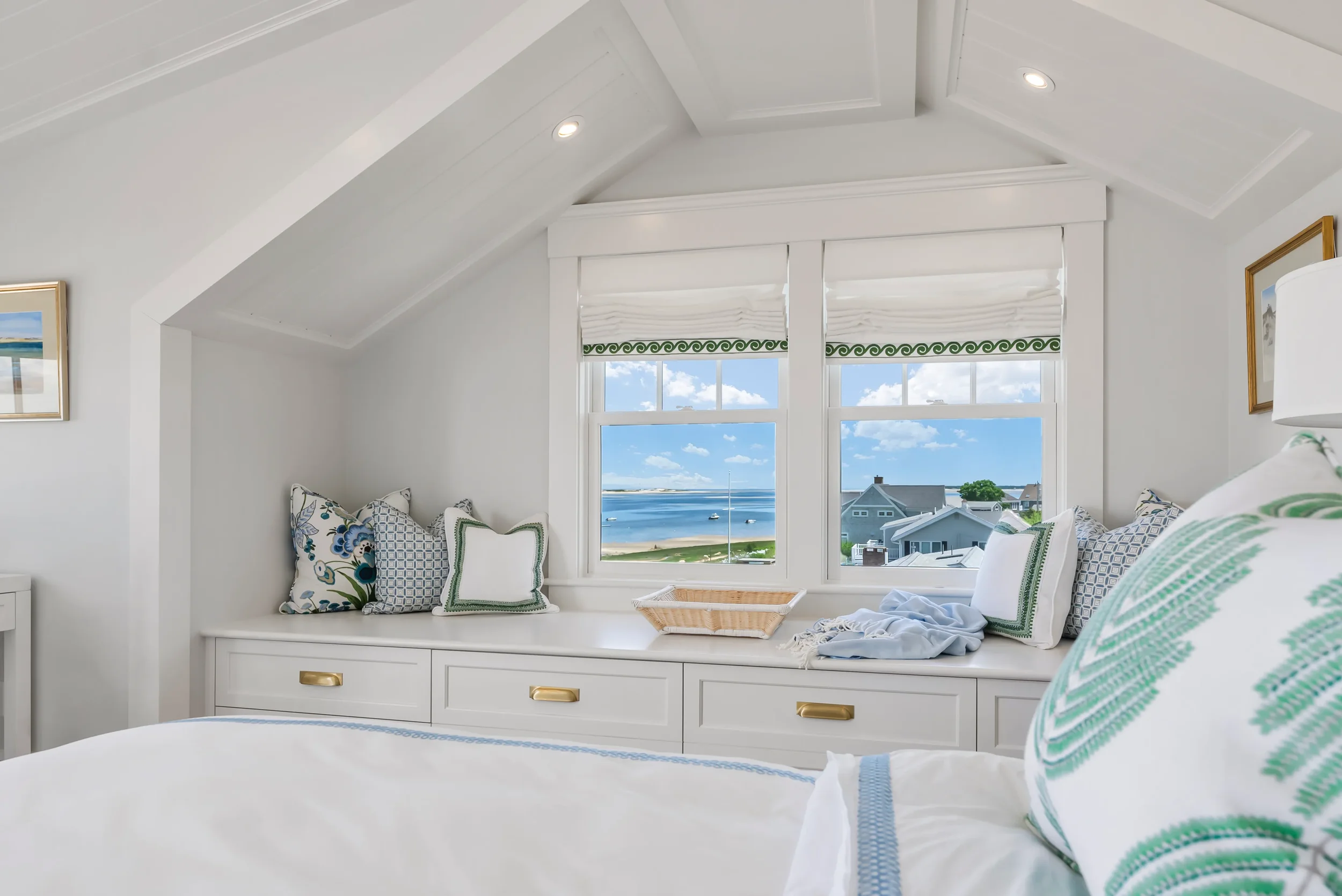 Bedroom with window seat, view of ocean and houses, white walls, bedding, decorative pillows, lamp, and art on wall