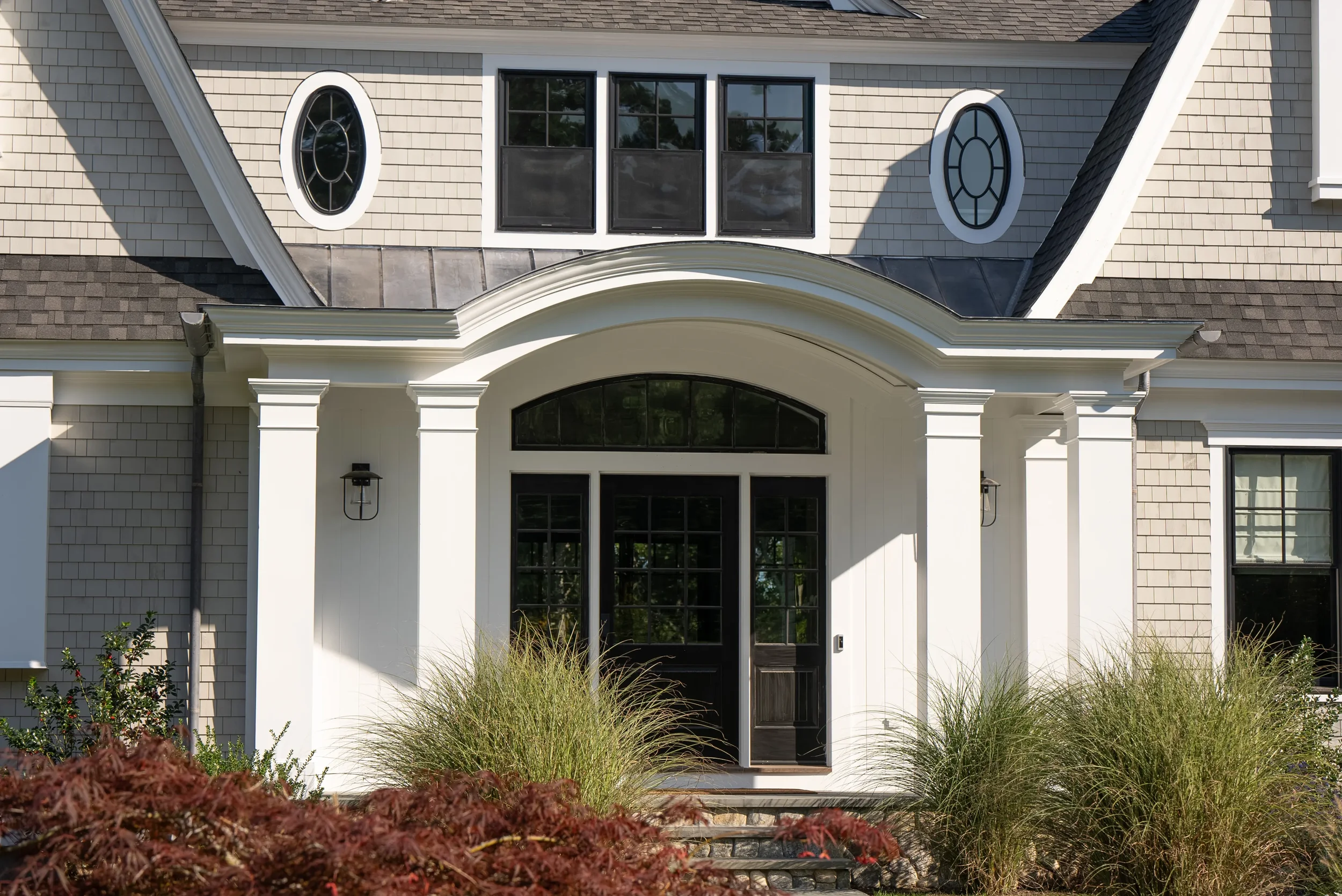 Front entrance of a house with a black door, white trim, and large windows, surrounded by tall ornamental grasses and shrubs.