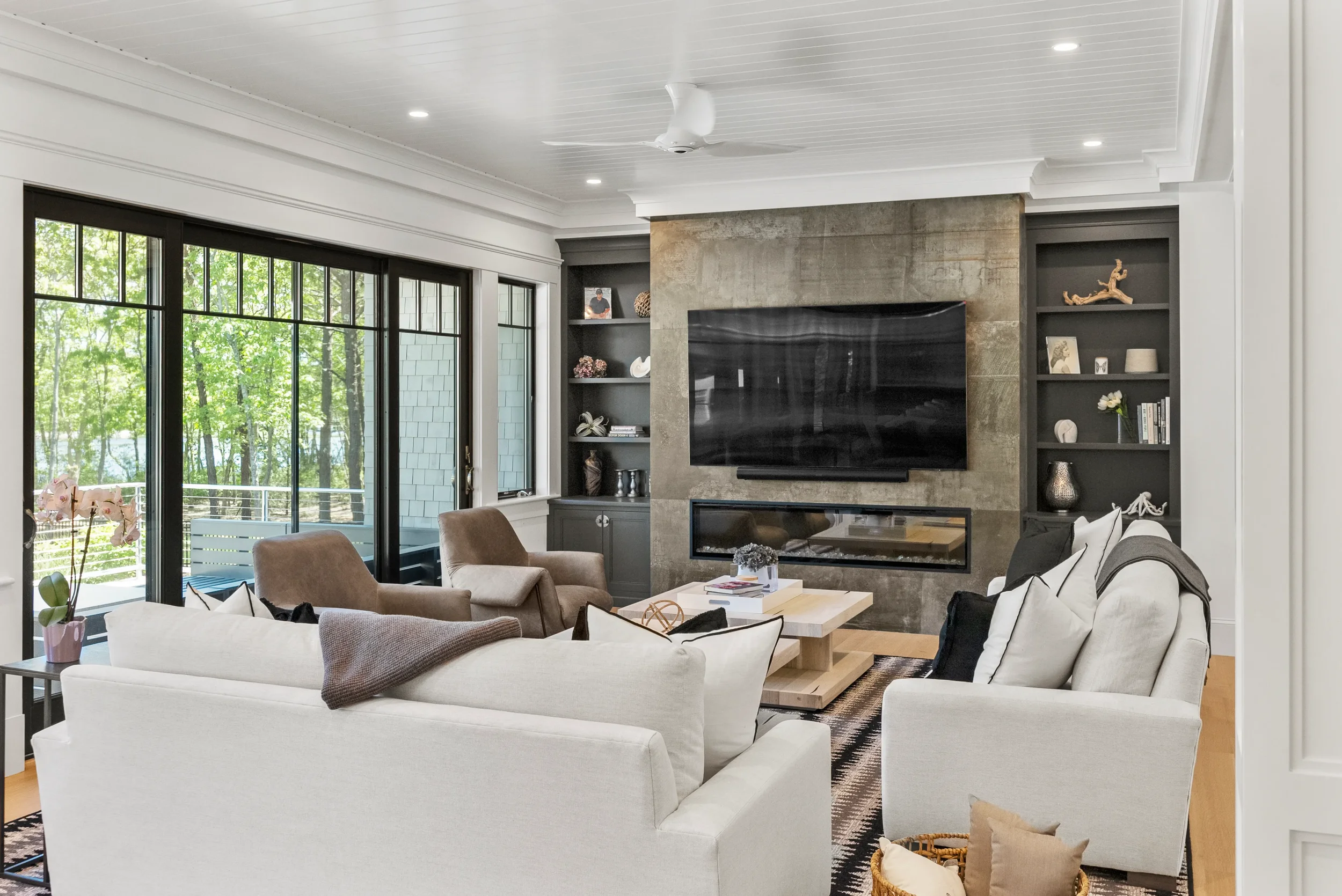 Living room with large windows, white walls, a ceiling fan, flat-screen TV on a stone wall, beige and white sofas, brown armchairs, a coffee table, decorative shelves, and a city view outside.