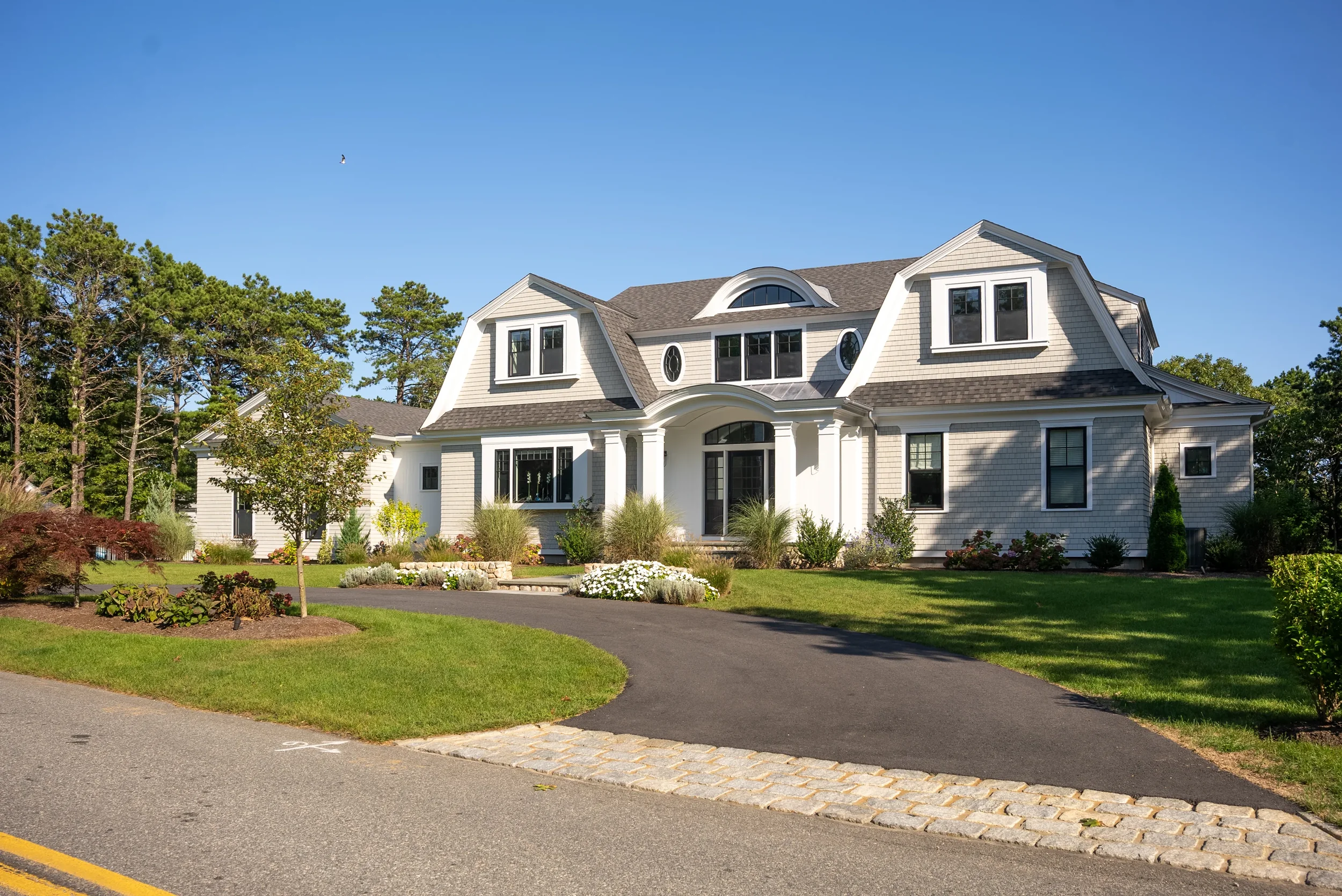 Large white house with grey roof, multiple windows, and a curved front porch, surrounded by a well-maintained lawn and garden, under a clear blue sky.