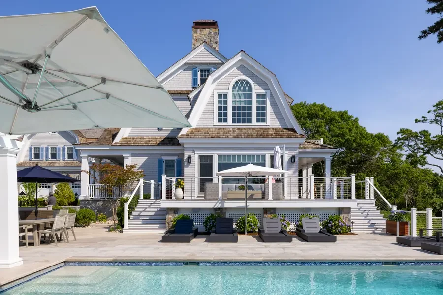 A luxurious house with a spacious backyard featuring a swimming pool, outdoor seating, sunny weather, and lush green trees.