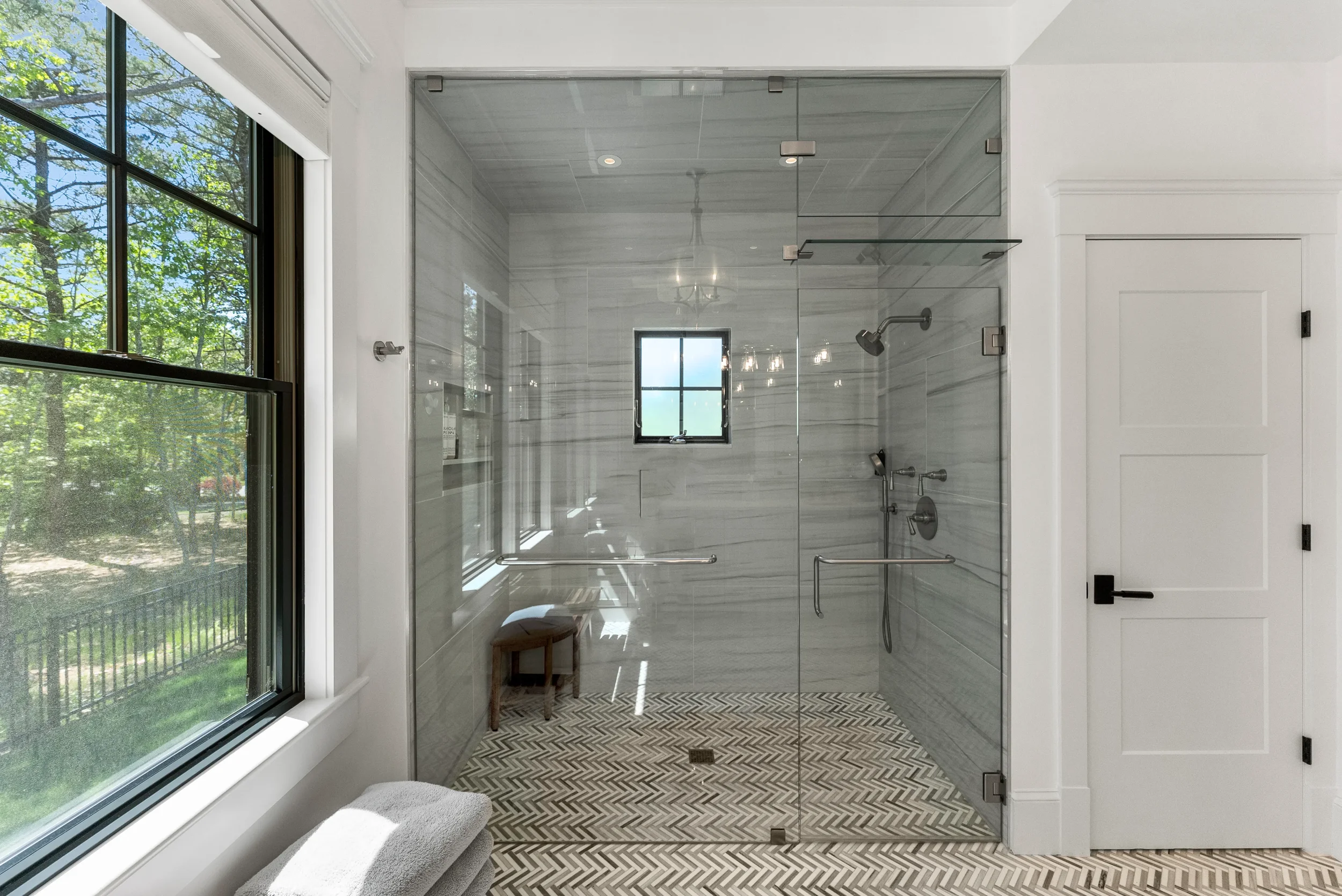 Modern bathroom with a glass-enclosed shower, white wood-paneled ceiling, large window showing green trees outside, patterned floor tiles, and a white door.
