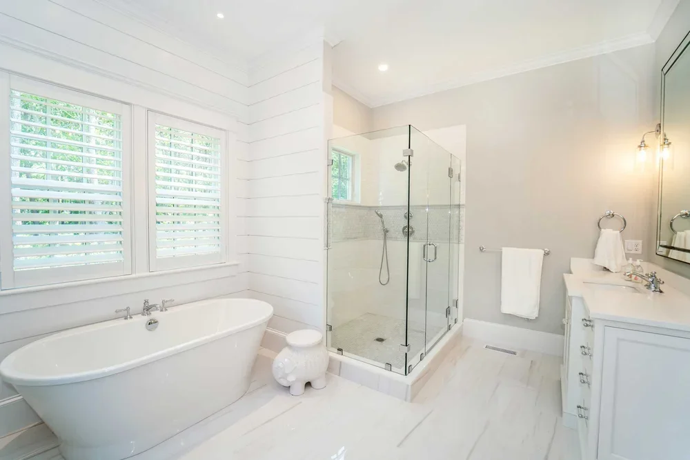 Bright bathroom featuring a white freestanding bathtub, a glass-enclosed shower with a small window, a white vanity with double sinks and a large mirror, and a towel mounted on the wall.