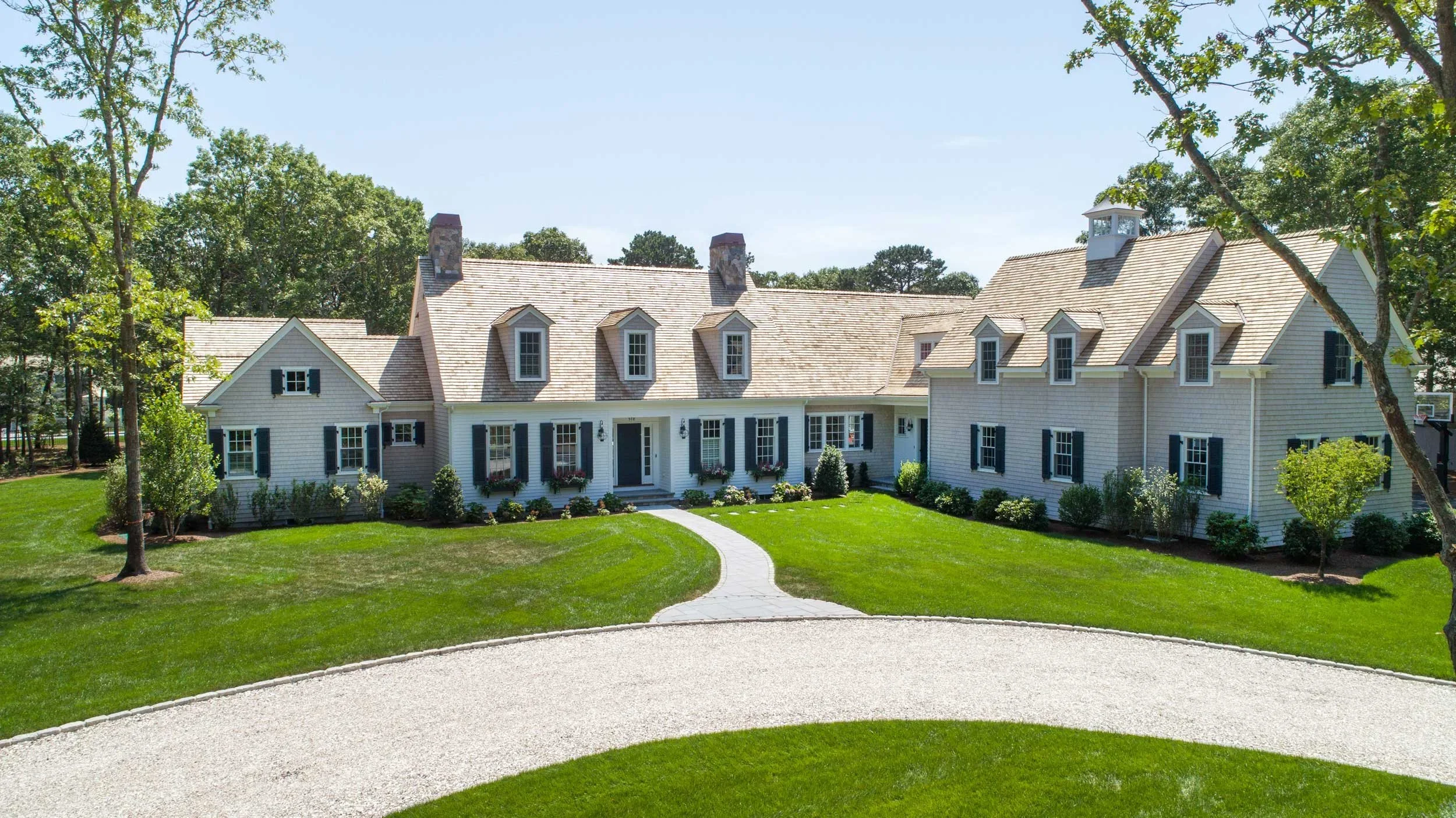 Large white house with black shutters, multiple dormer windows, surrounded by green lawn and trees, under a clear blue sky.