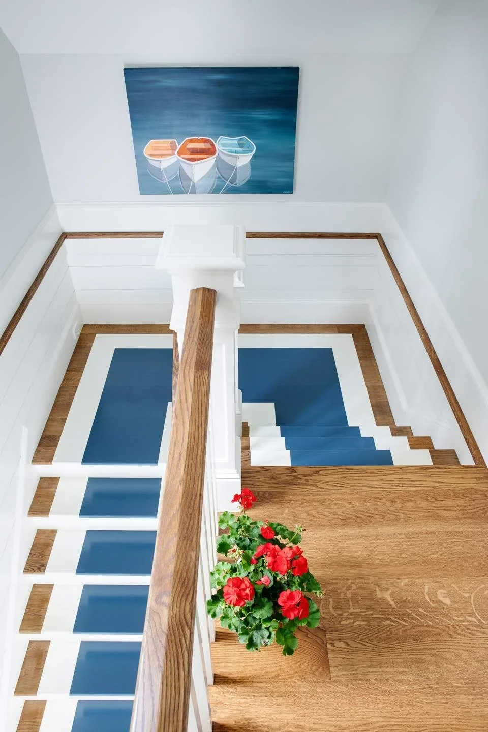 Top view of a staircase with blue and white carpet, a wooden handrail, a red potted flower, and a painting of three colorful boats on water mounted on a white wall.
