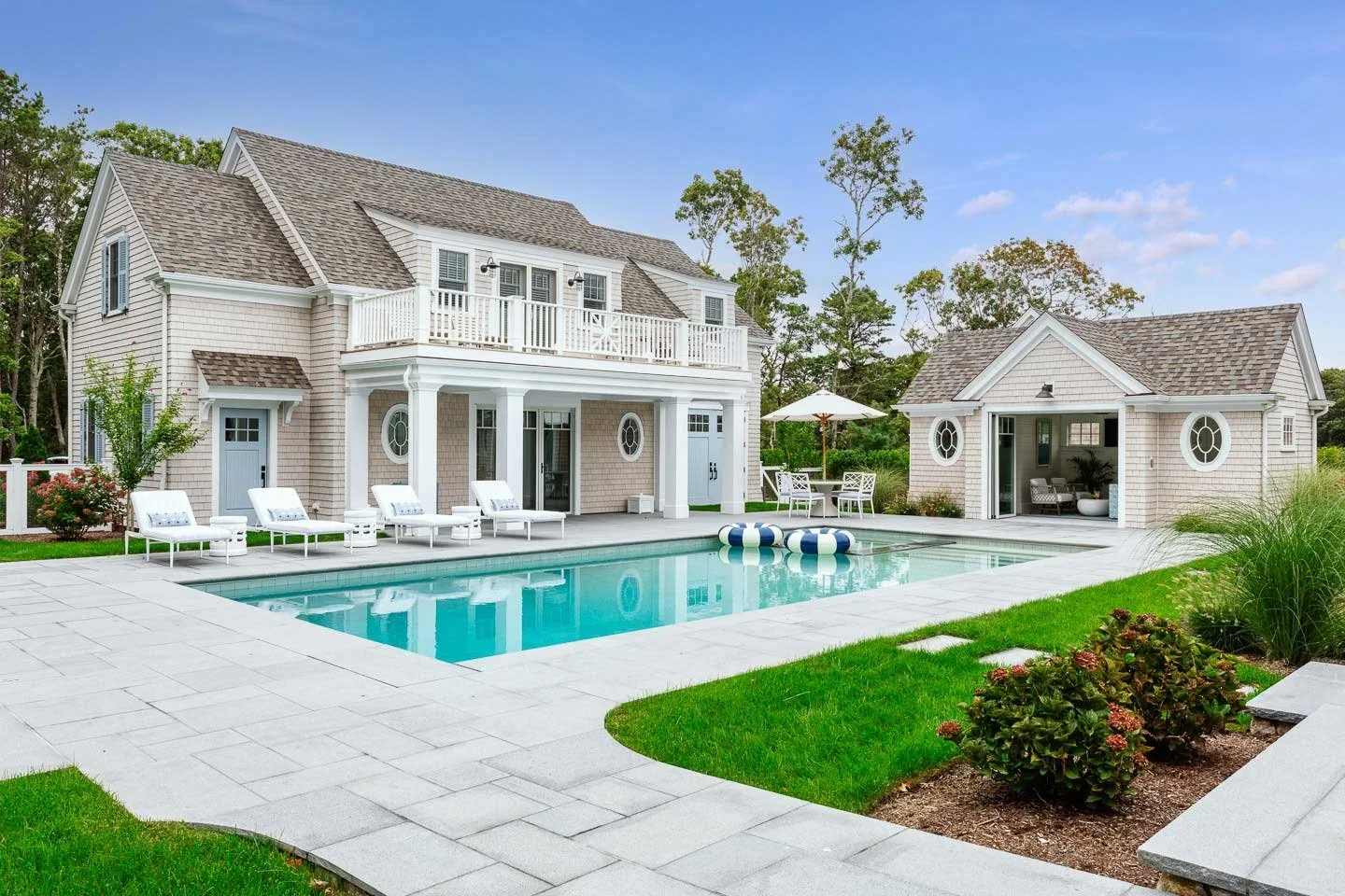 This image showcases a luxurious residential backyard designed for relaxation and upscale outdoor living. At the center is a large swimming pool, surrounded by a meticulously maintained stone patio and lush green lawn. Four white lounge chairs line t