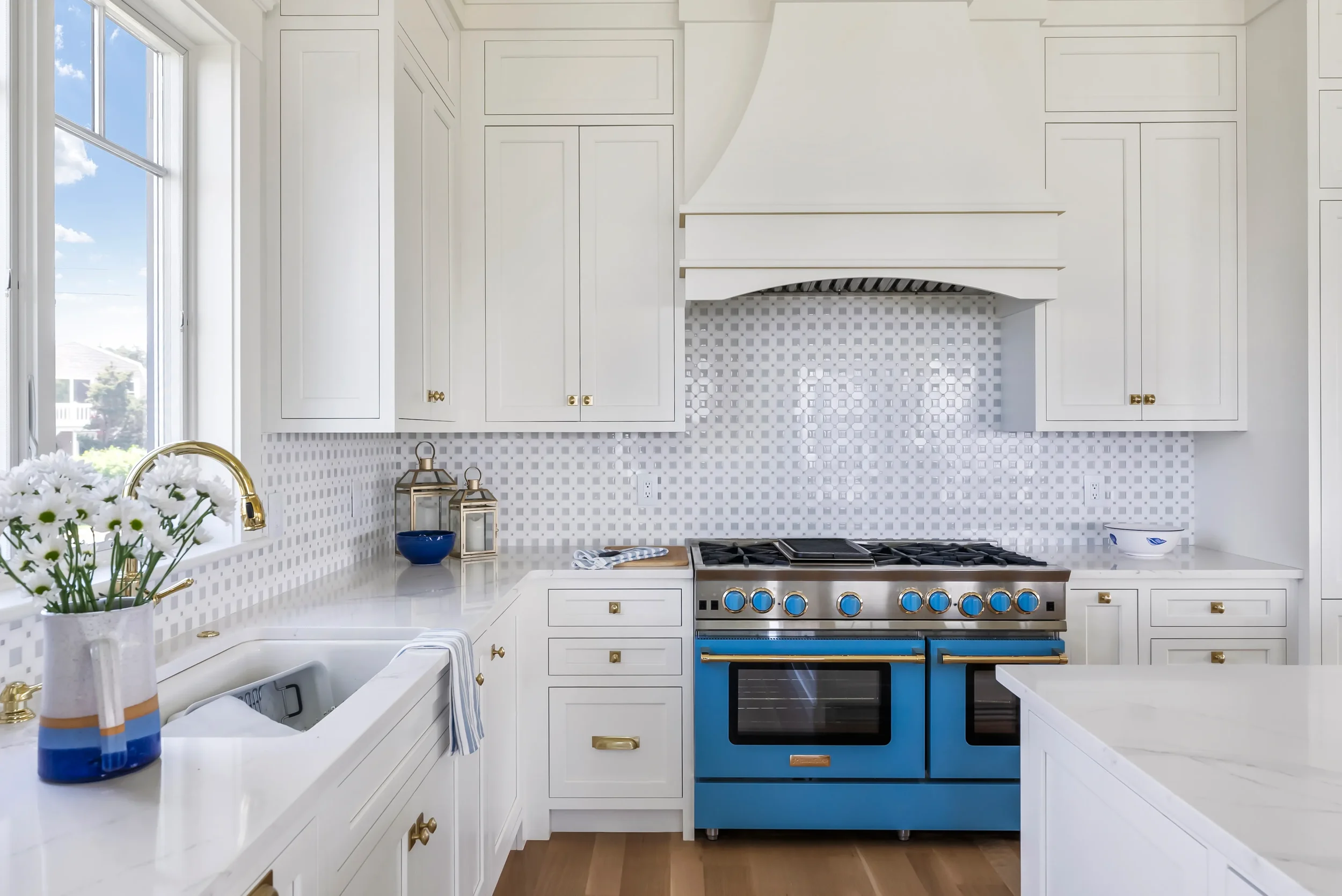 A bright, modern kitchen with white cabinets, gold hardware, a white marble countertop, and a blue stove. There are decorative lanterns, a vase with white flowers, and a window with a view of a sunny neighborhood.