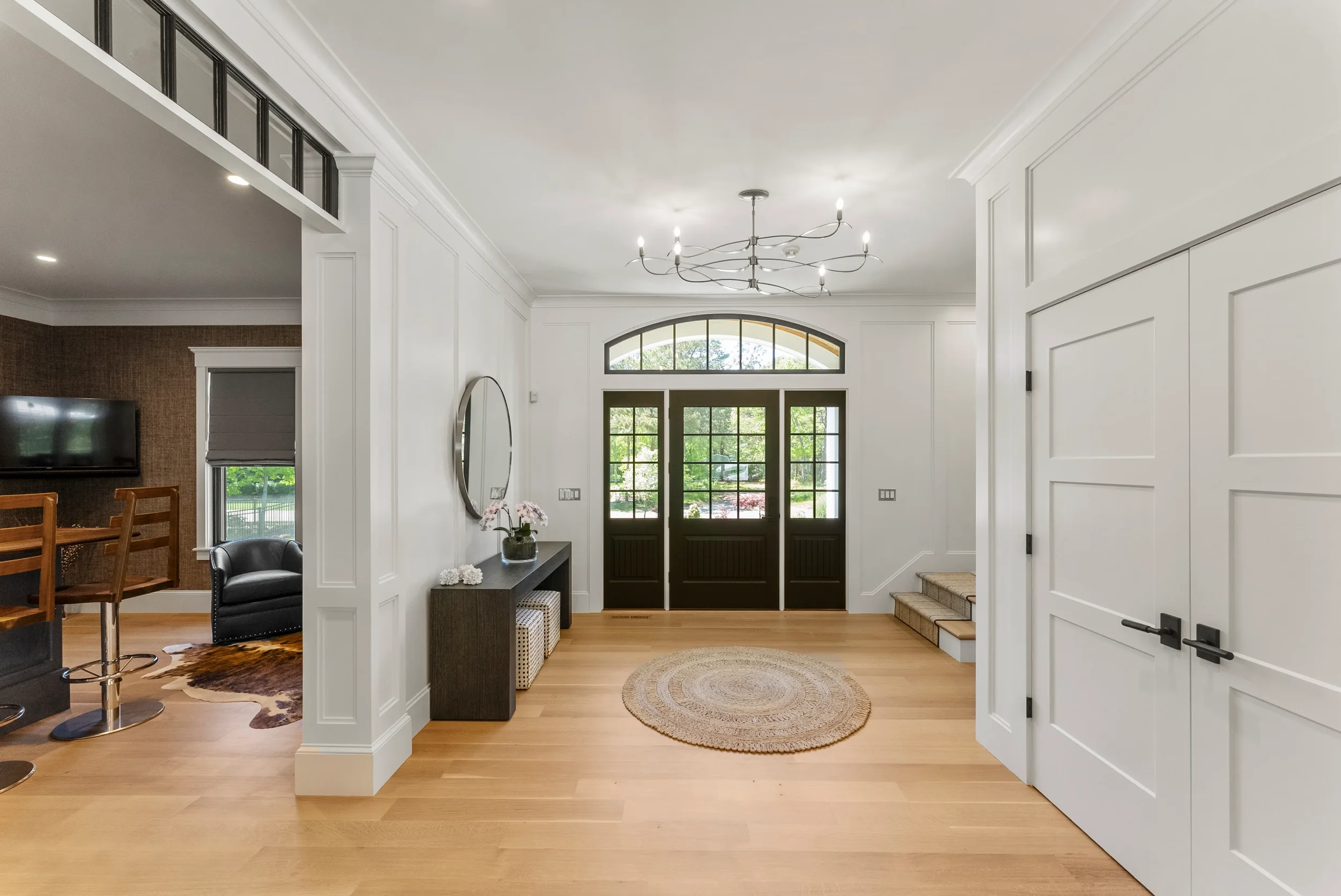 Bright and spacious entryway with light wood floors, white walls, and a black front door with glass panes. A circular woven rug is placed in the center, and a modern chandelier hangs from the ceiling. To the left, a hallway leads to a living area wit
