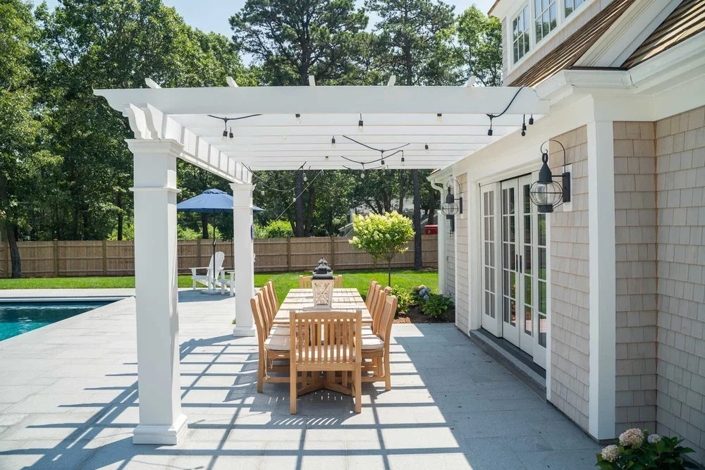 An outdoor patio area with a wooden dining table and chairs, a pergola with string lights, a pool in the background, and a house with double glass doors.