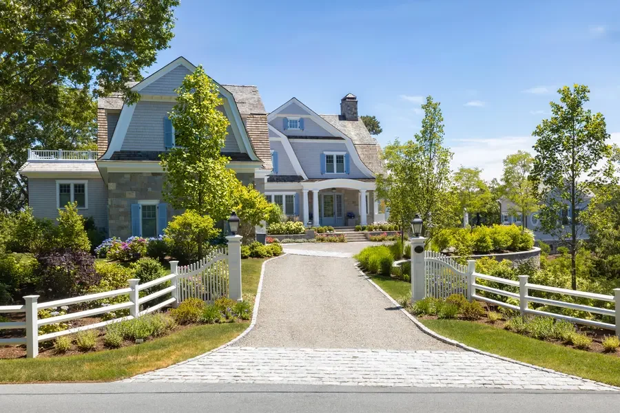 A large, elegant house with a stone and white siding exterior, surrounded by well-maintained landscaping, trees, and a white picket fence, with a paved driveway leading up to the front entrance.