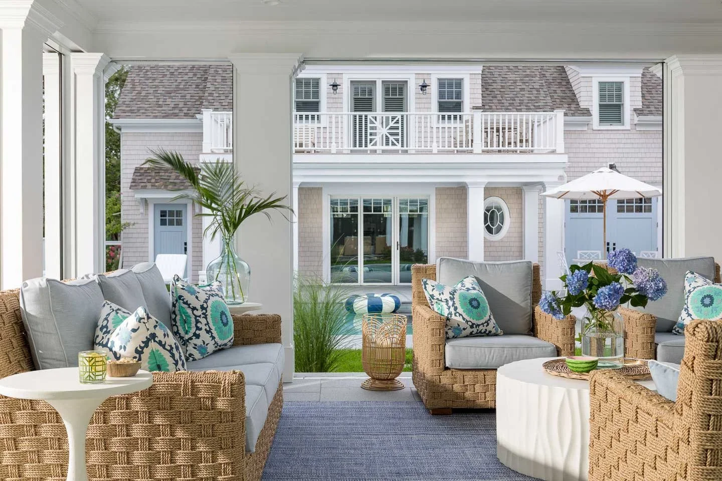 Sunlit indoor porch with wicker furniture, patterned cushions, and a view of a large house with an upper balcony outside.
