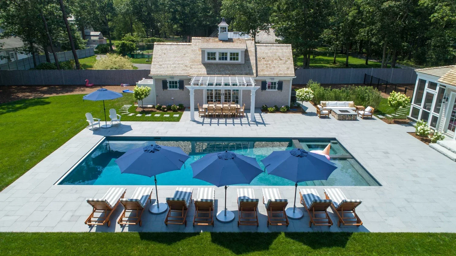 A backyard with a swimming pool, four blue umbrellas, six lounge chairs with striped cushions, a patio with a dining table and chairs, a shed, and a house with a porch, surrounded by green grass and trees.