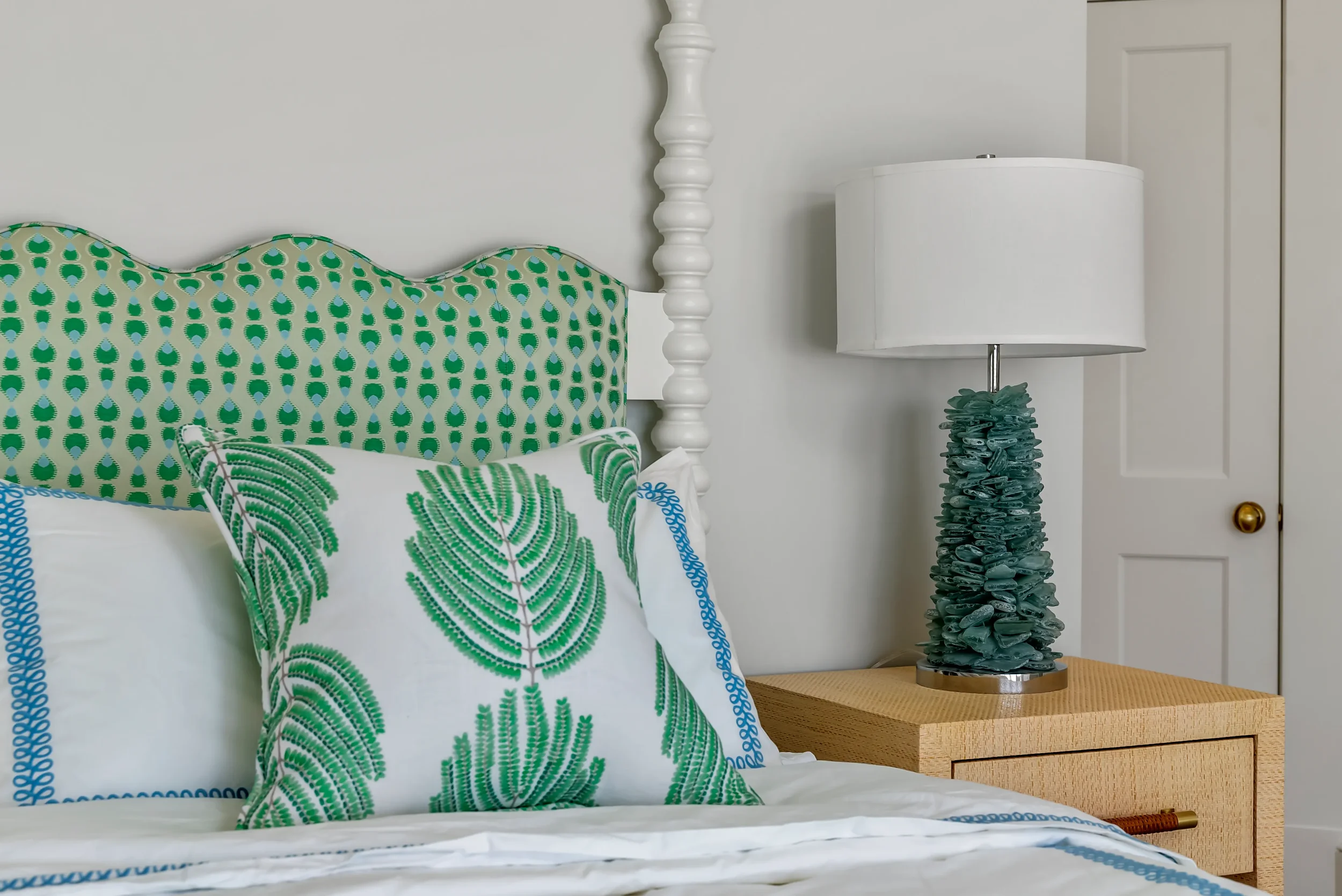 A bedroom with a green patterned headboard, two decorative pillows with green leaf designs, a wooden nightstand, and a table lamp with a textured base and a white lampshade.
