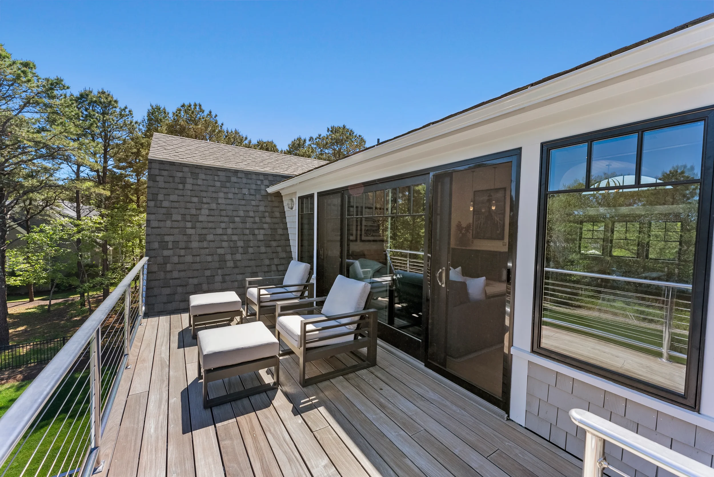 Outdoor balcony with wooden flooring, modern furniture including chairs and ottomans, surrounded by a metal railing, overlooking trees and neighboring houses on a clear, sunny day.