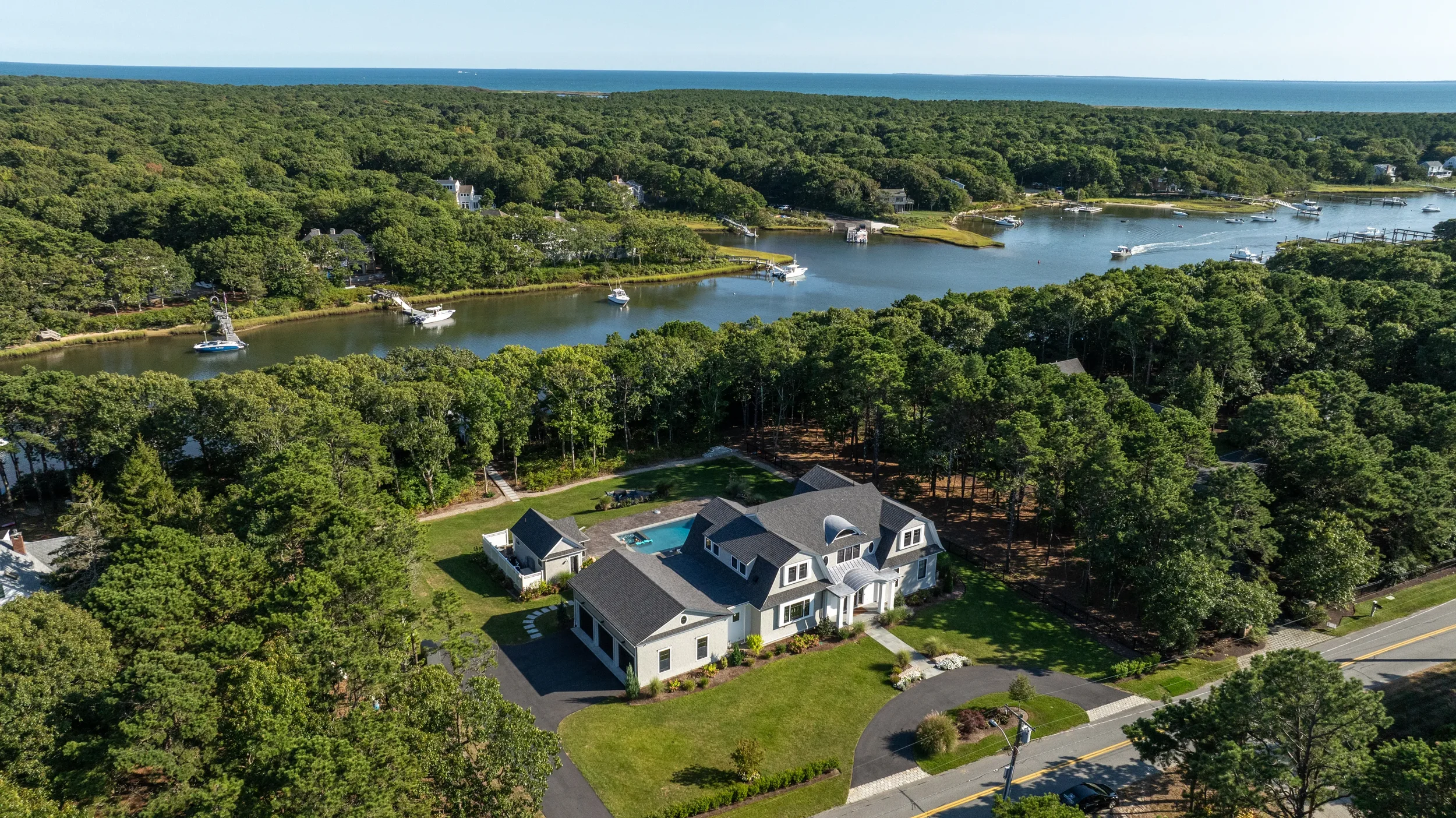 A large house with a backyard and swimming pool, surrounded by trees, overlooking a waterway with boats and houses along the shoreline, with a distant view of the ocean.
