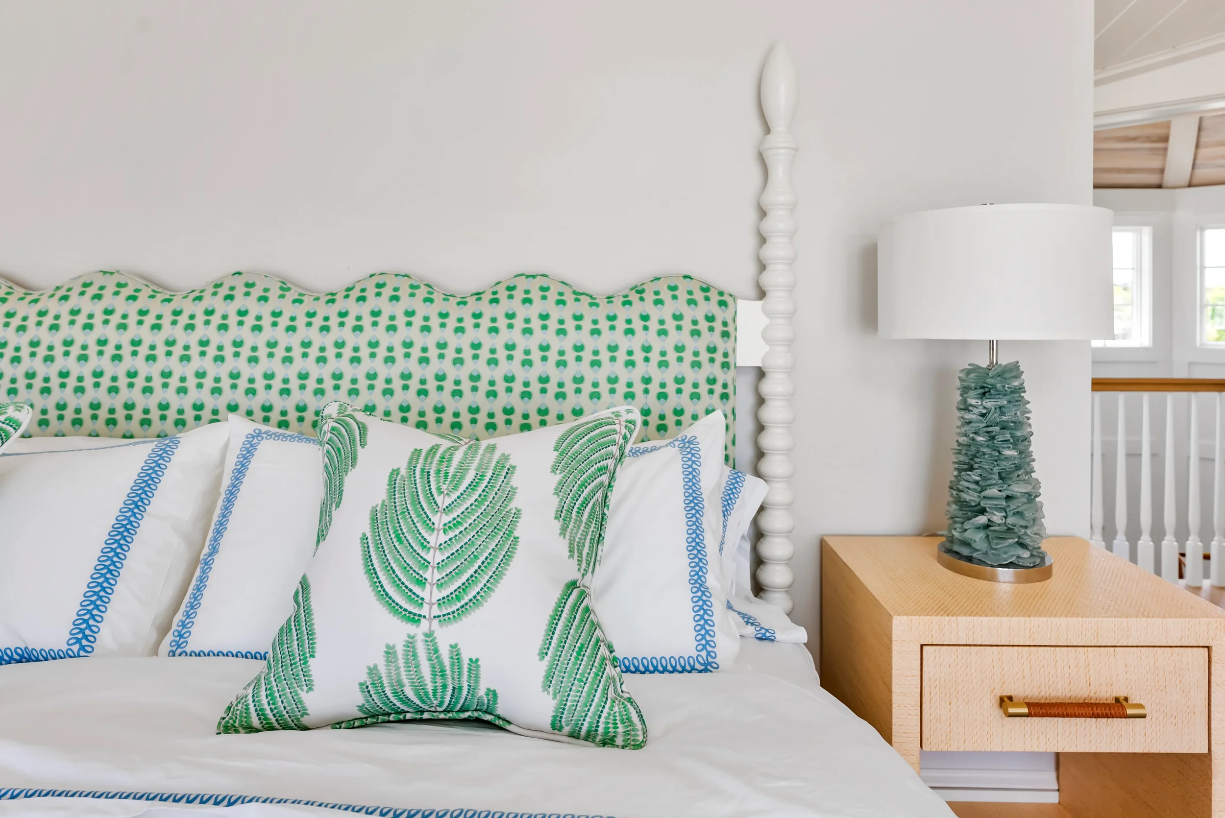 A bedroom with a green and white patterned headboard, white pillows with green and blue leaf designs, a wooden nightstand with a textured turquoise bug, and a large white lamp. The room has a white wall and windows in the background.