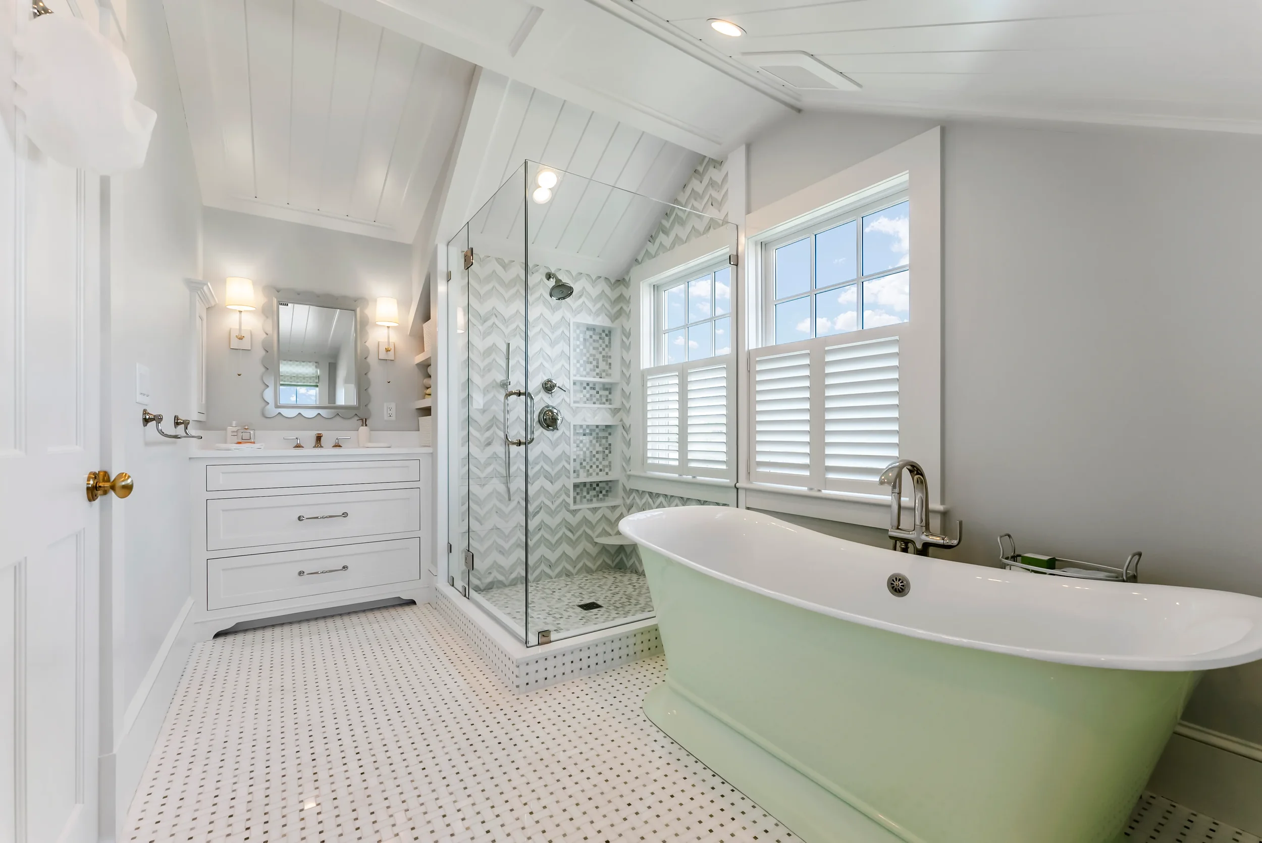 Bright bathroom with a freestanding bathtub, glass-enclosed shower with patterned tile, white vanity with mirror, and large windows with white shutters.