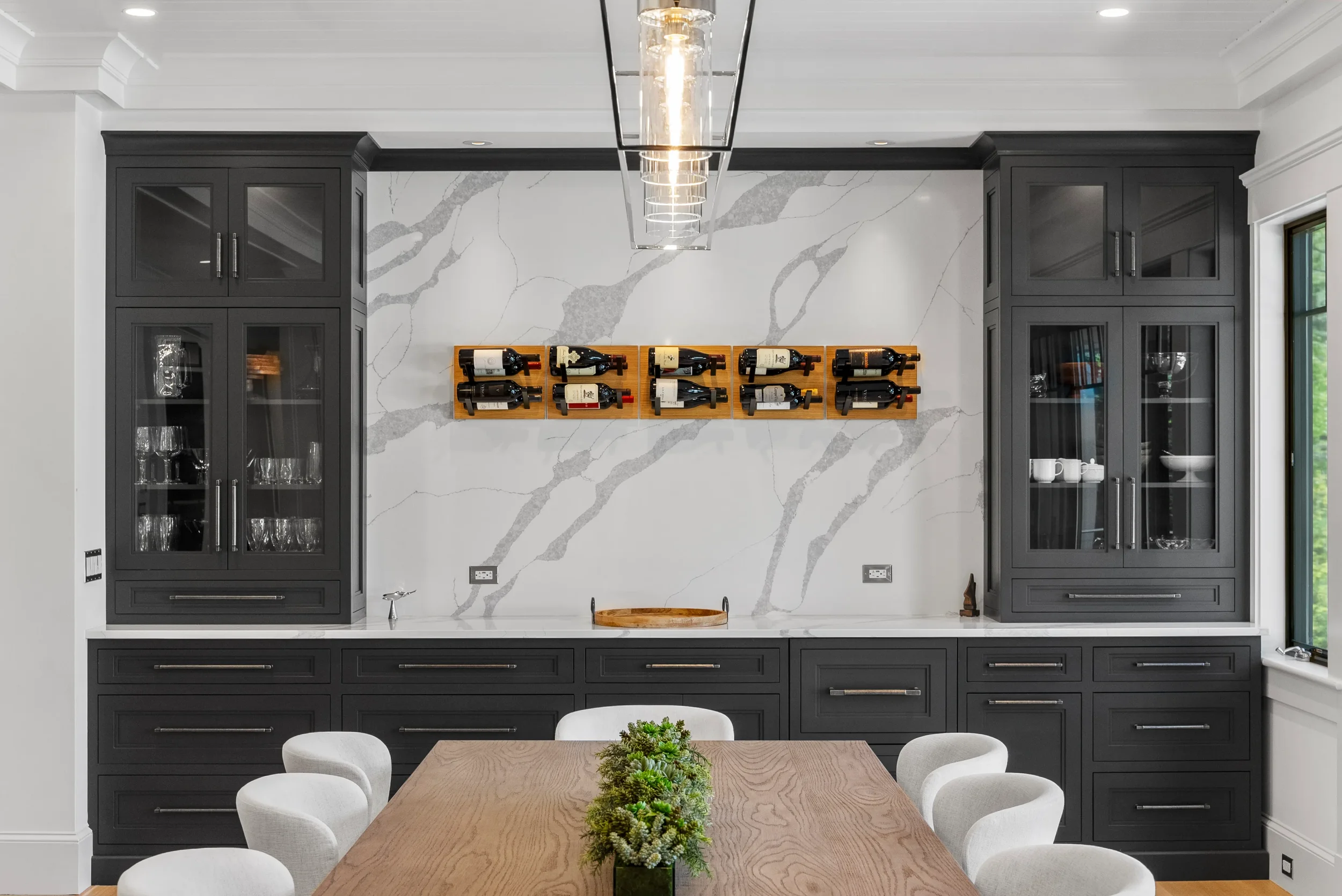 Modern dining room with black cabinetry, a white marble backsplash with gray veining, wine bottles on a wall-mounted rack, and a wooden dining table with white upholstered chairs.