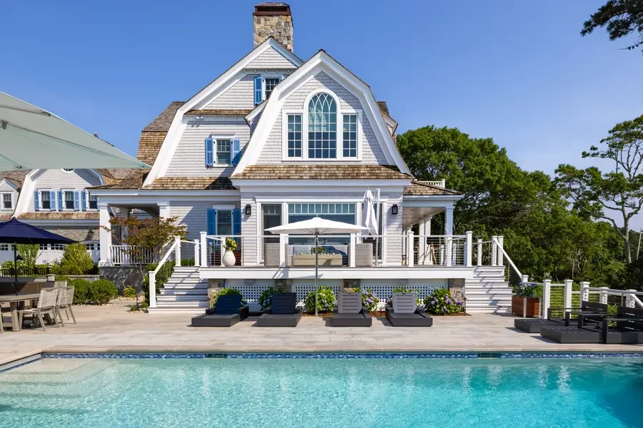 A large white house with a deck, patio furniture, a swimming pool in the foreground, and lush green trees in the background under a clear blue sky.