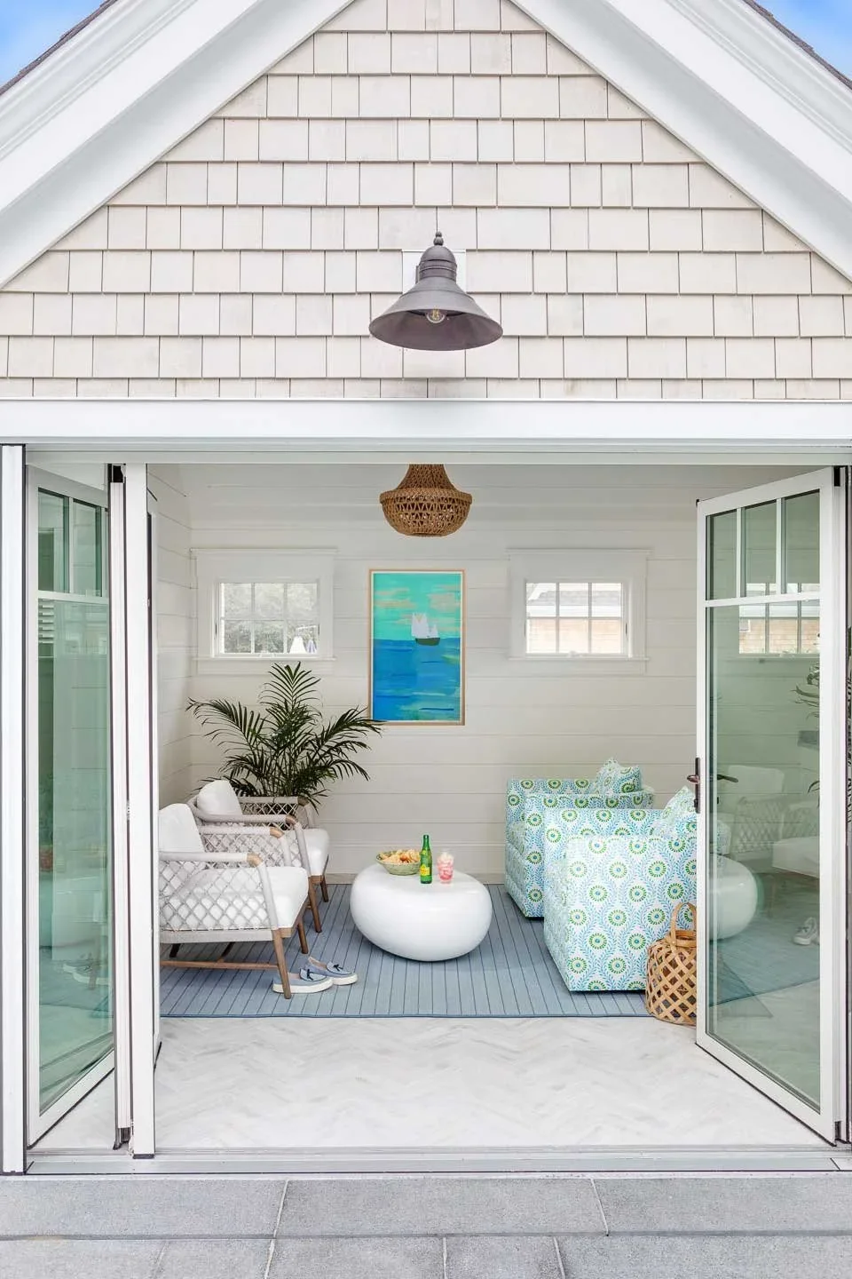 A cozy indoor patio with white walls, two windows, a framed painting of a sailboat on the water, three patterned armchairs, a white round coffee table with drinks, a potted plant, and a hanging wicker light fixture. It opens to an outdoor space with