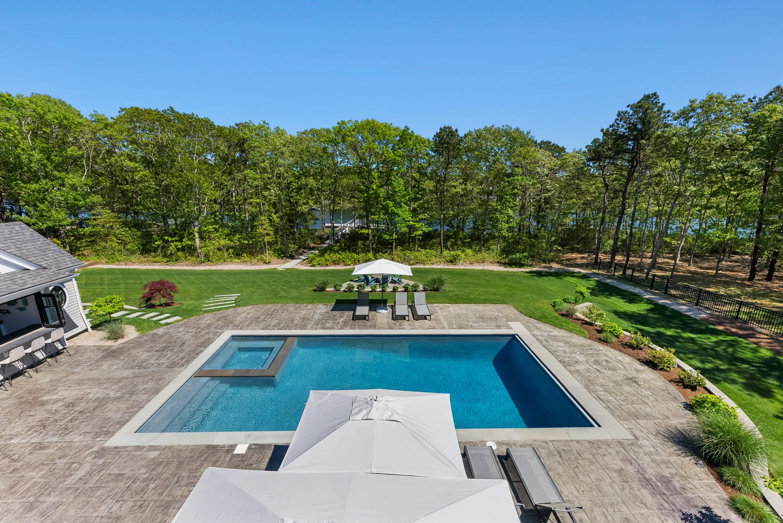A backyard with a swimming pool, lounge chairs, a large white umbrella, a lush green lawn, trees, and a glimpse of a lake in the background.