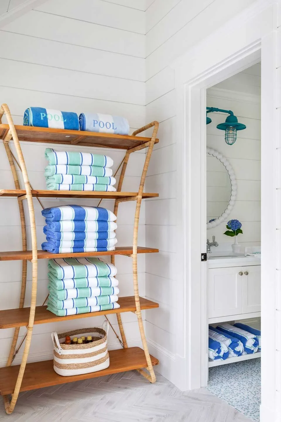 Stacked towels in shades of blue, white, and green on wooden shelves, with a glimpse of a bathroom featuring a sink, mirror, and flowers to the right.