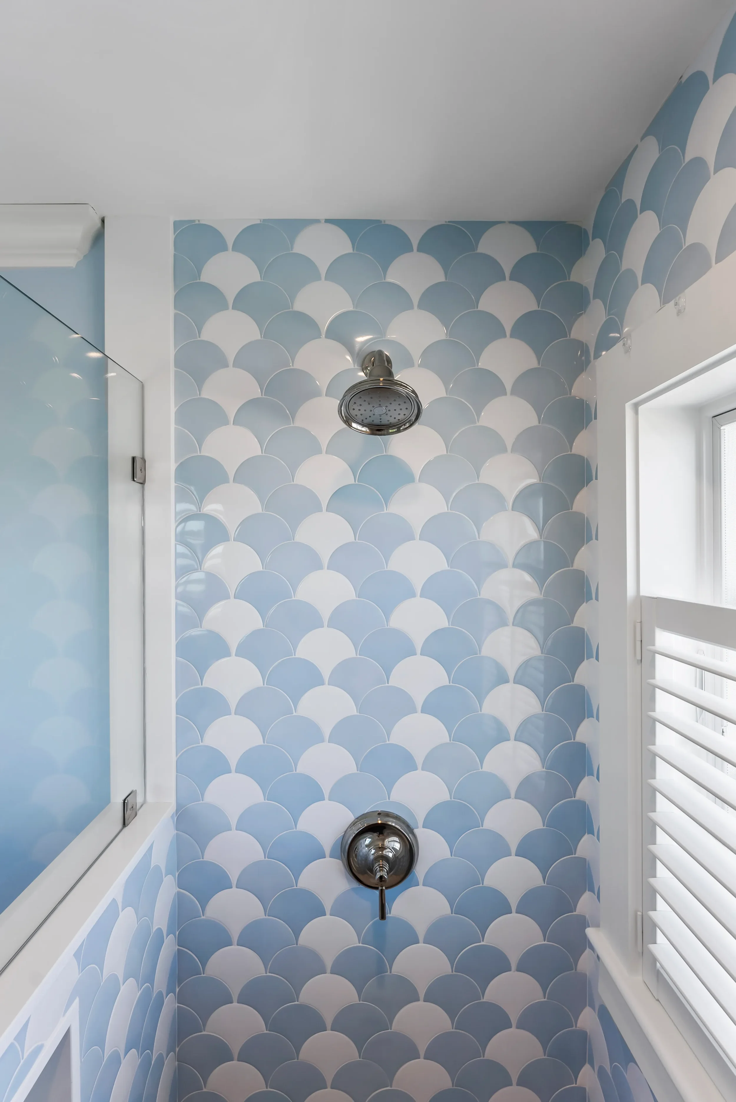 Shower area with patterned blue and white scalloped tiles, a silver shower head, and a window with white shutters.