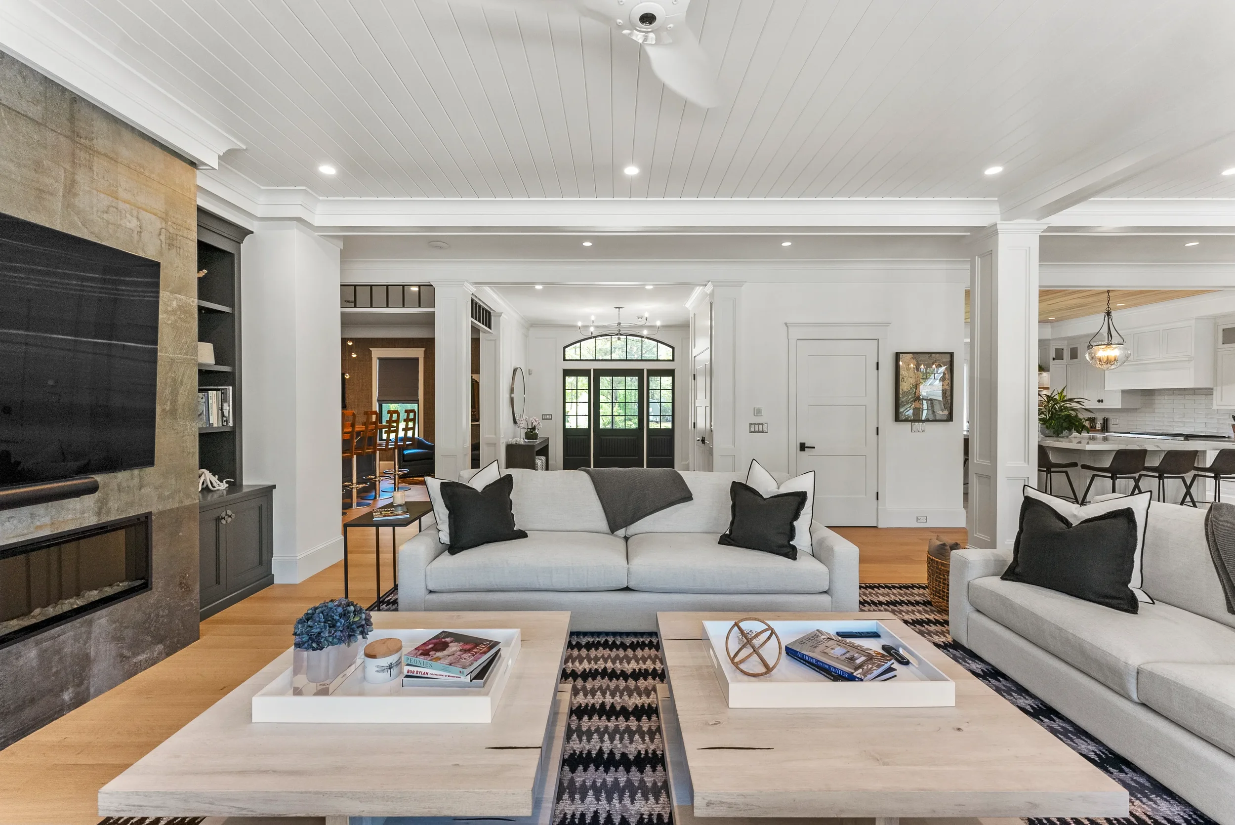 Living room with white sofas, black and white pillows, a light wood coffee table, and a fireplace with a flat-screen TV above. The room has white walls, a beamed ceiling, and hardwood floors. The background shows a hallway with a front door and a gli