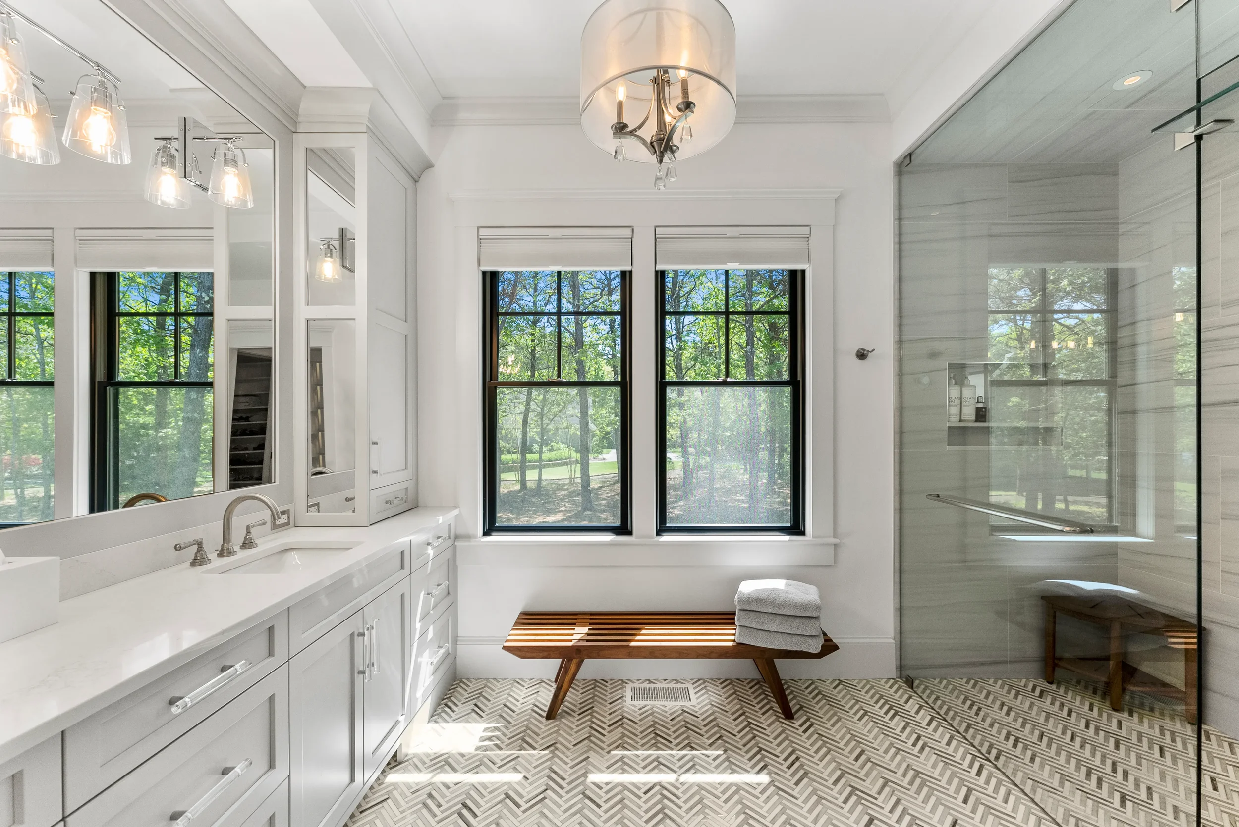 Bright, modern bathroom with white cabinets, a large mirror, and a white countertop. Two large windows with black frames reveal trees outside. A wooden bench with folded towels is beneath the windows. A glass-enclosed shower area is visible on the ri