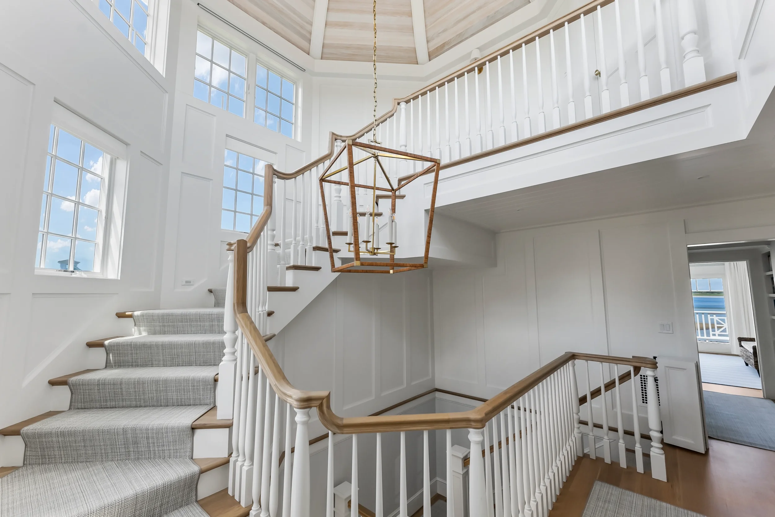 Interior view of a staircase and landing in a bright, modern house with white walls, large windows, and a wooden chandelier.