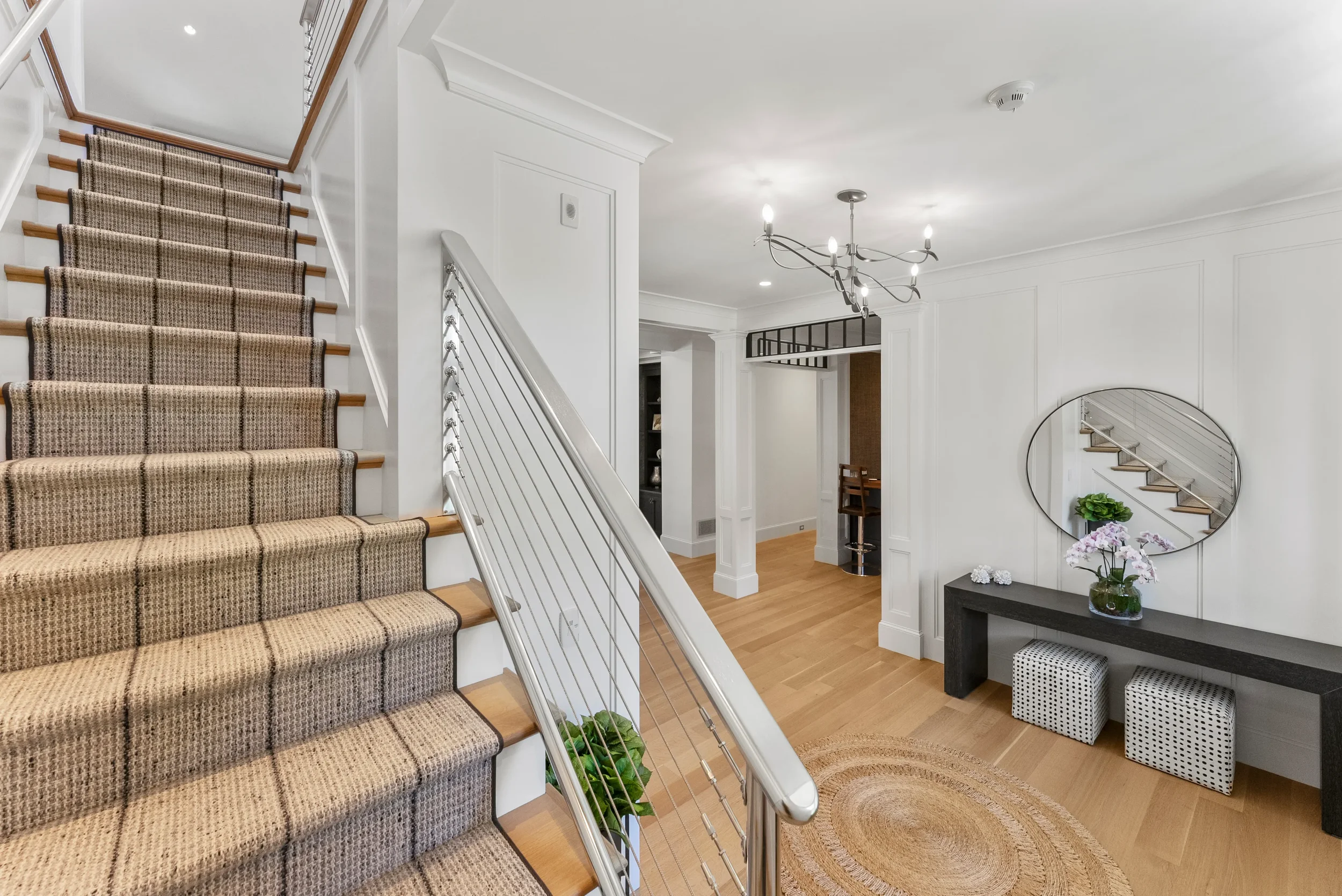 Interior of a modern home with a staircase, white walls, wooden floors, a black console table with a vase of flowers, wall mirror, and contemporary decor.