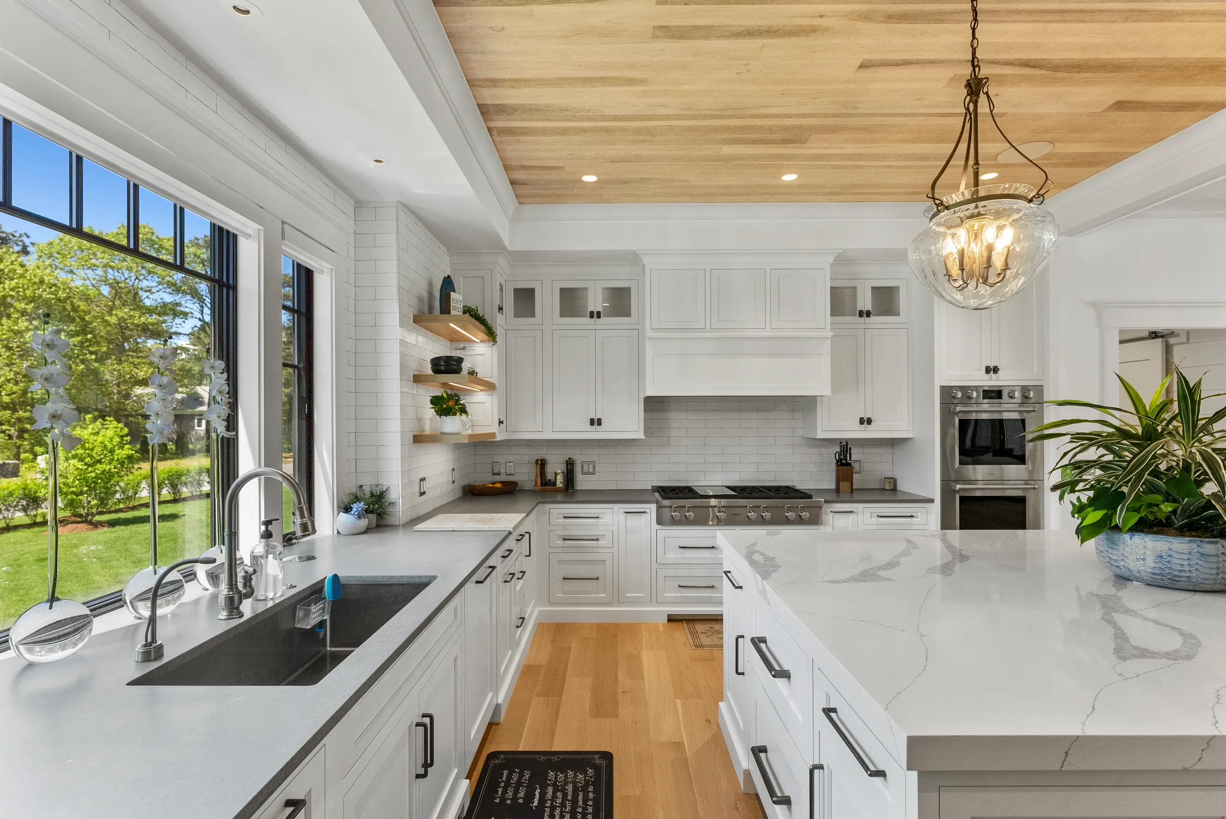 Bright kitchen with white cabinets, marble countertops, large window showing outdoor greenery, wooden ceiling, chandelier, and a plant on the island.