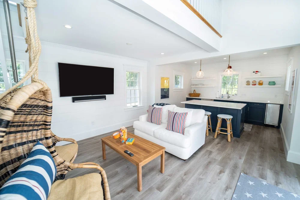 Open-concept living room and kitchen with white walls, wooden floors, and modern furnishings, featuring a hanging swing chair, a white sofa with striped pillows, a wooden coffee table, and a black kitchen island with stools.