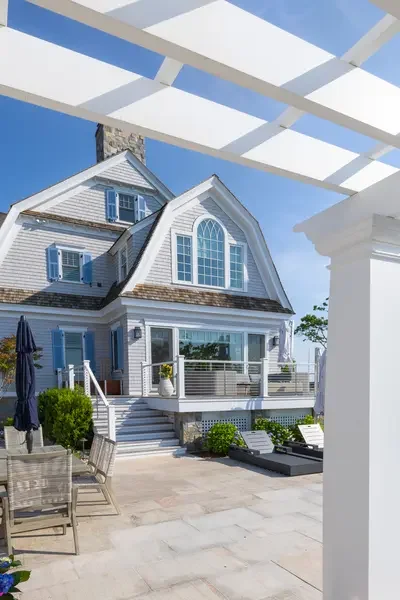 White beach house with multiple stories, large windows, and a patio with outdoor dining furniture, under a white pergola.