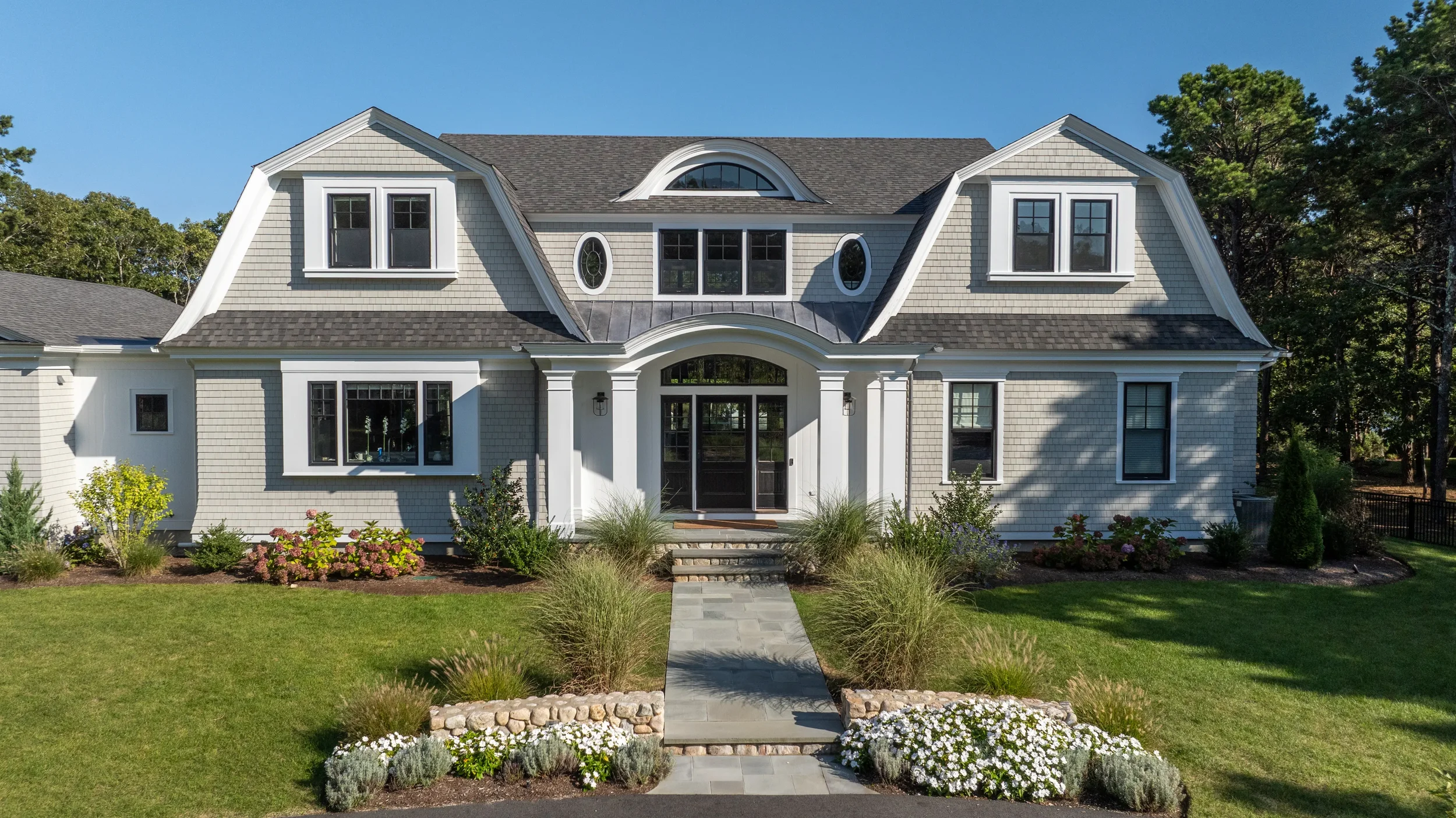 front view of a large, two-story house with gray shingles, white trim, and a landscaped yard with a stone walkway, grass, and flowering bushes.