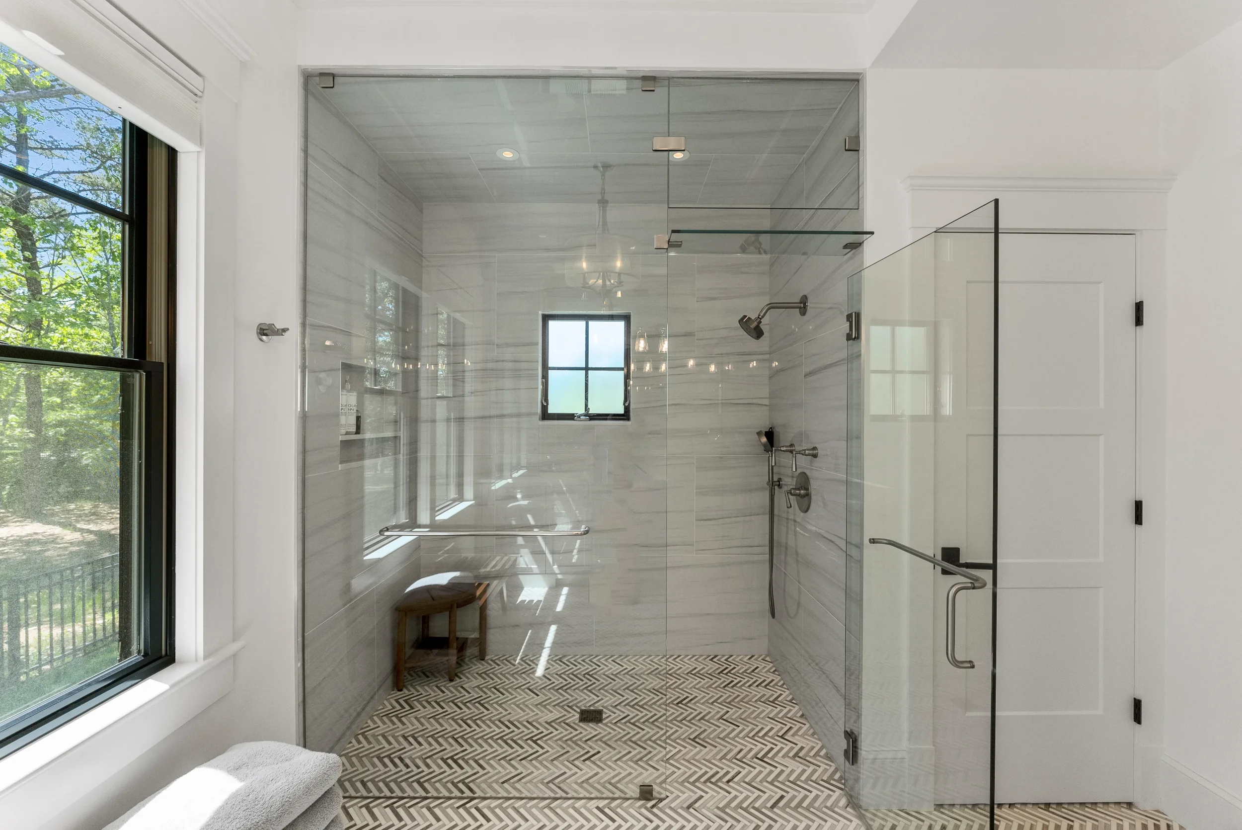 Modern walk-in shower with glass walls, striped marble tiles, a small window, and a bench inside. The shower has a rainfall showerhead and a handheld spray, with a towel and window seat outside.